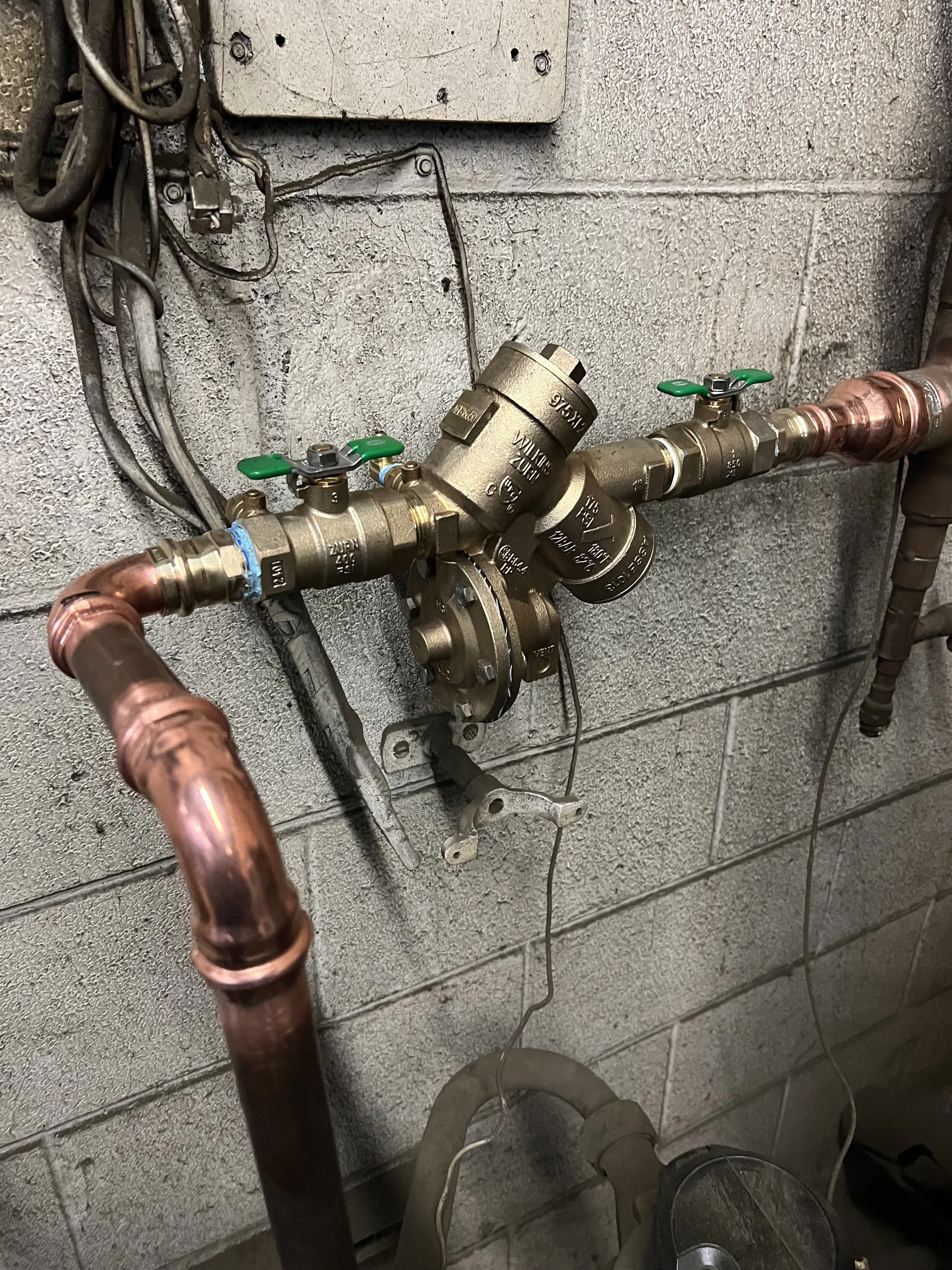 A plumbing system with copper pipes, brass valves, and a pressure regulator mounted on a cinder block wall.