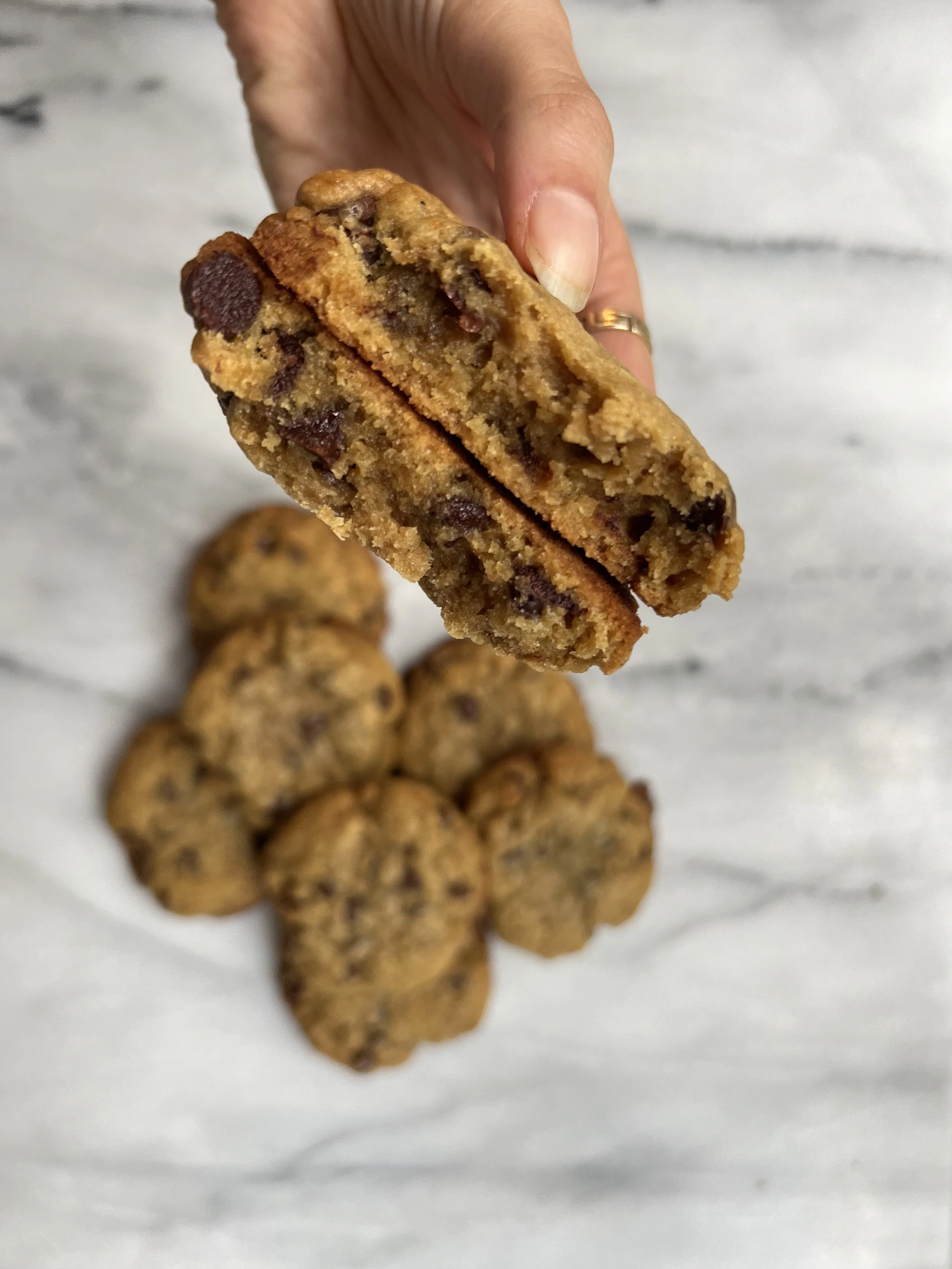 A hand holding a chocolate chip cookie cut in half, showing the inside with chocolate chips, with several whole cookies on a white, marbled surface in the background.