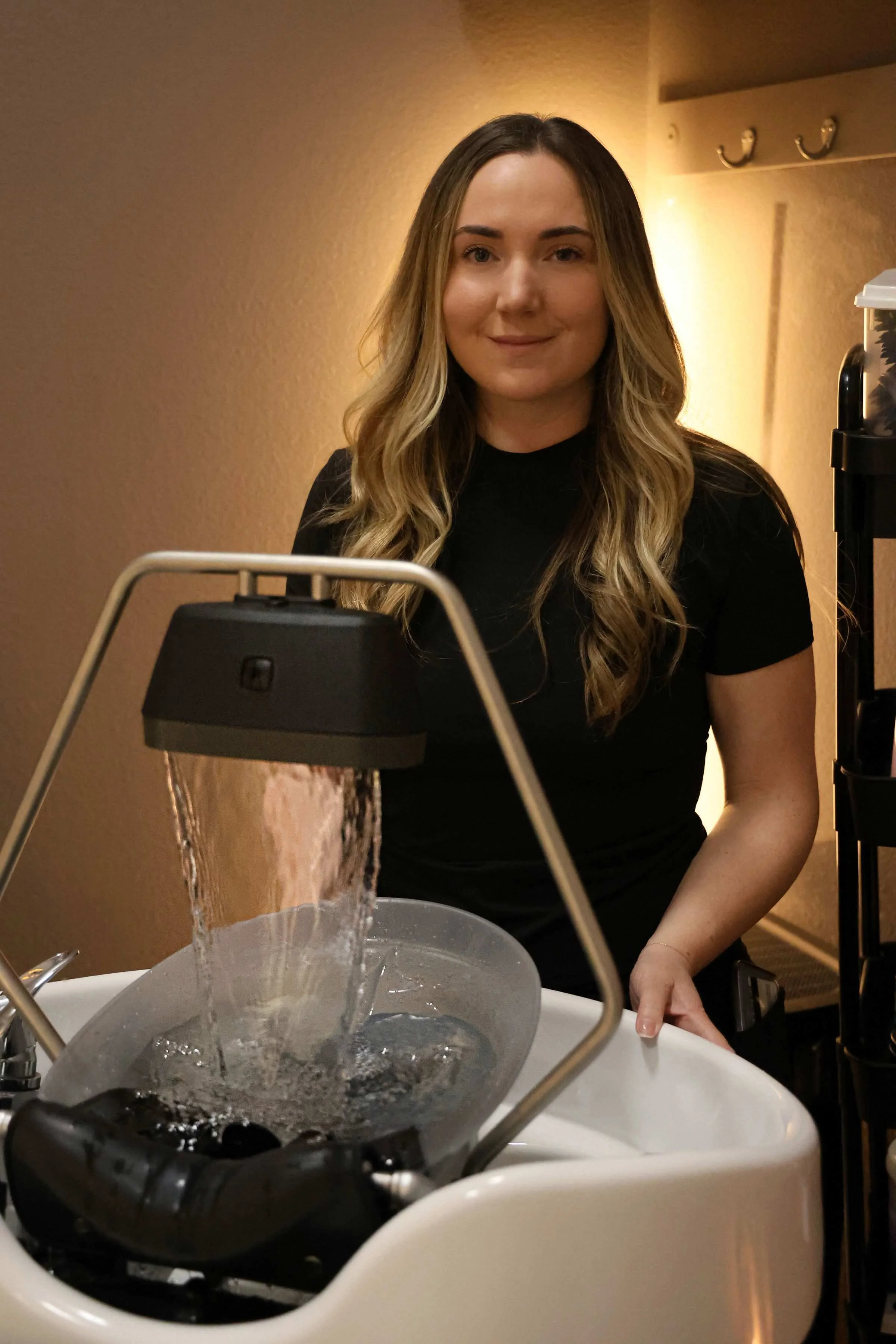 A woman with long, wavy blonde hair standing behind a sink with water flowing from a fountain, smiling at the camera, in a room with beige walls and a shelf on the right.