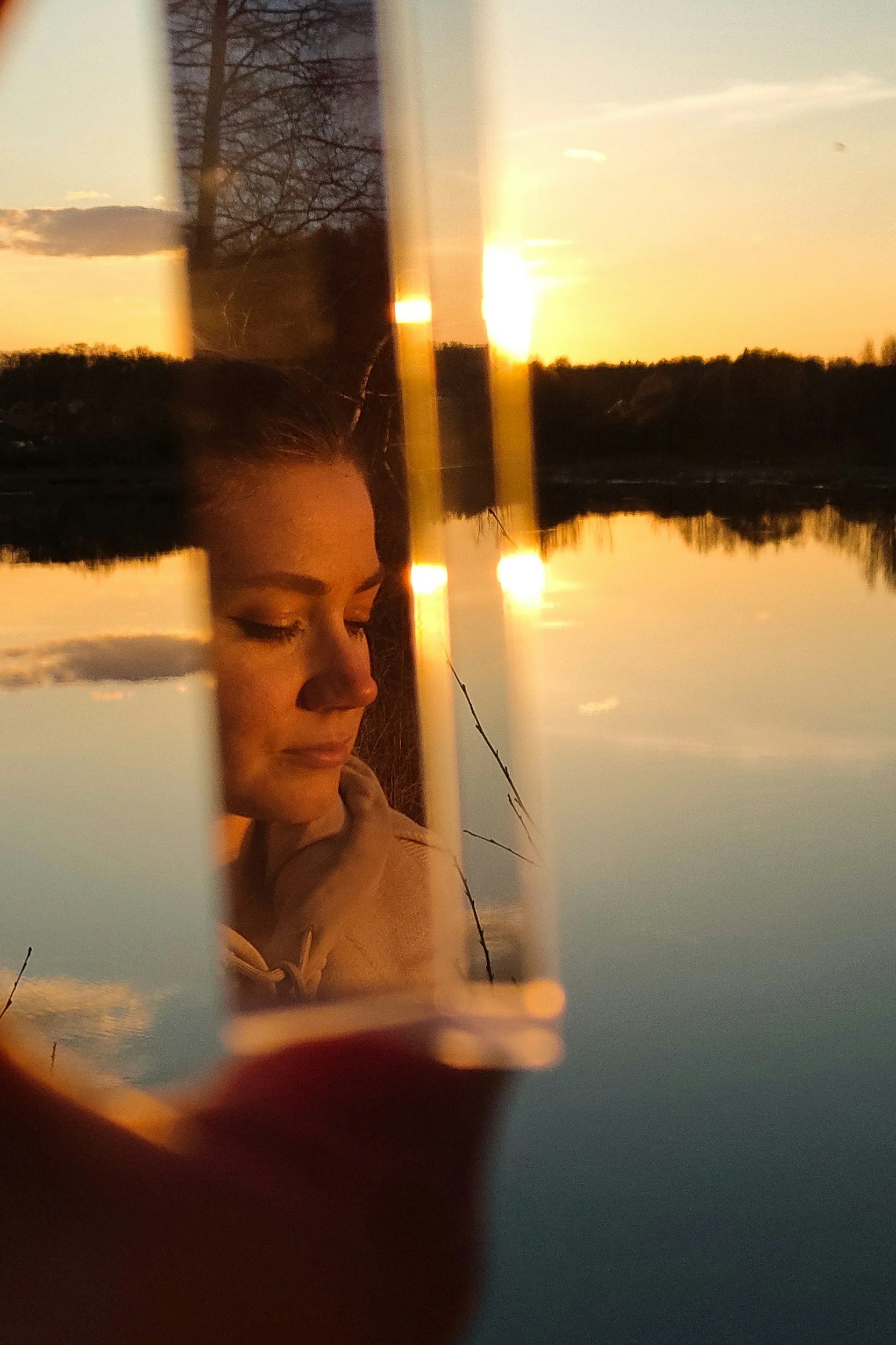 Reflection of a young woman with closed eyes, resting her chin on her hand, seen through a broken mirror reflecting a sunset over a lake with trees in the distance.
