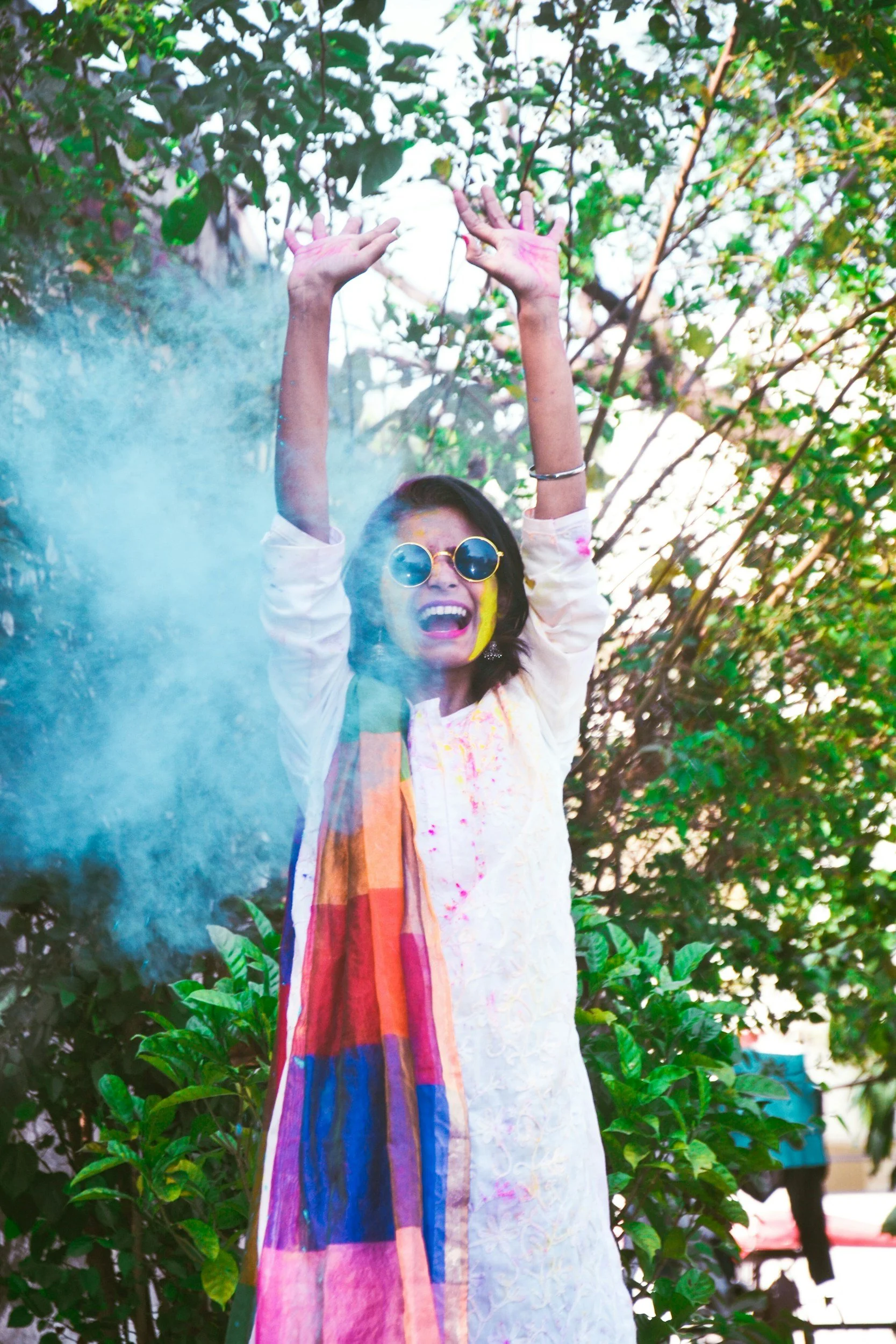 A woman wearing sunglasses and traditional Indian attire celebrating during Holi, covered in colorful powder with smoke around her, outdoors with green foliage in the background.