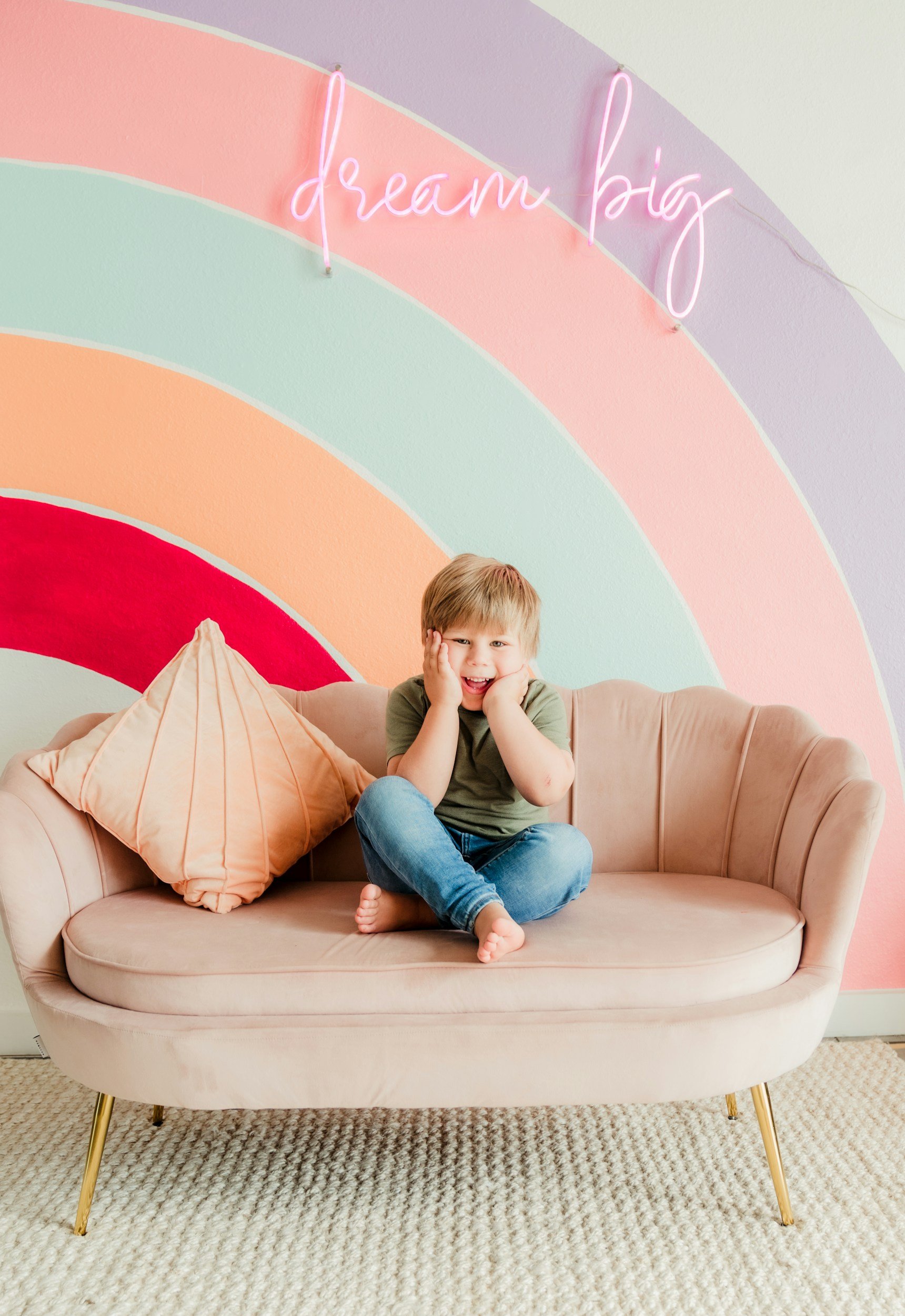 A young boy sitting on a pink velvet sofa with a coral pillow, smiling and holding his face with his hands, in front of a colorful rainbow painted wall with a pink neon sign that says 'dream big'.