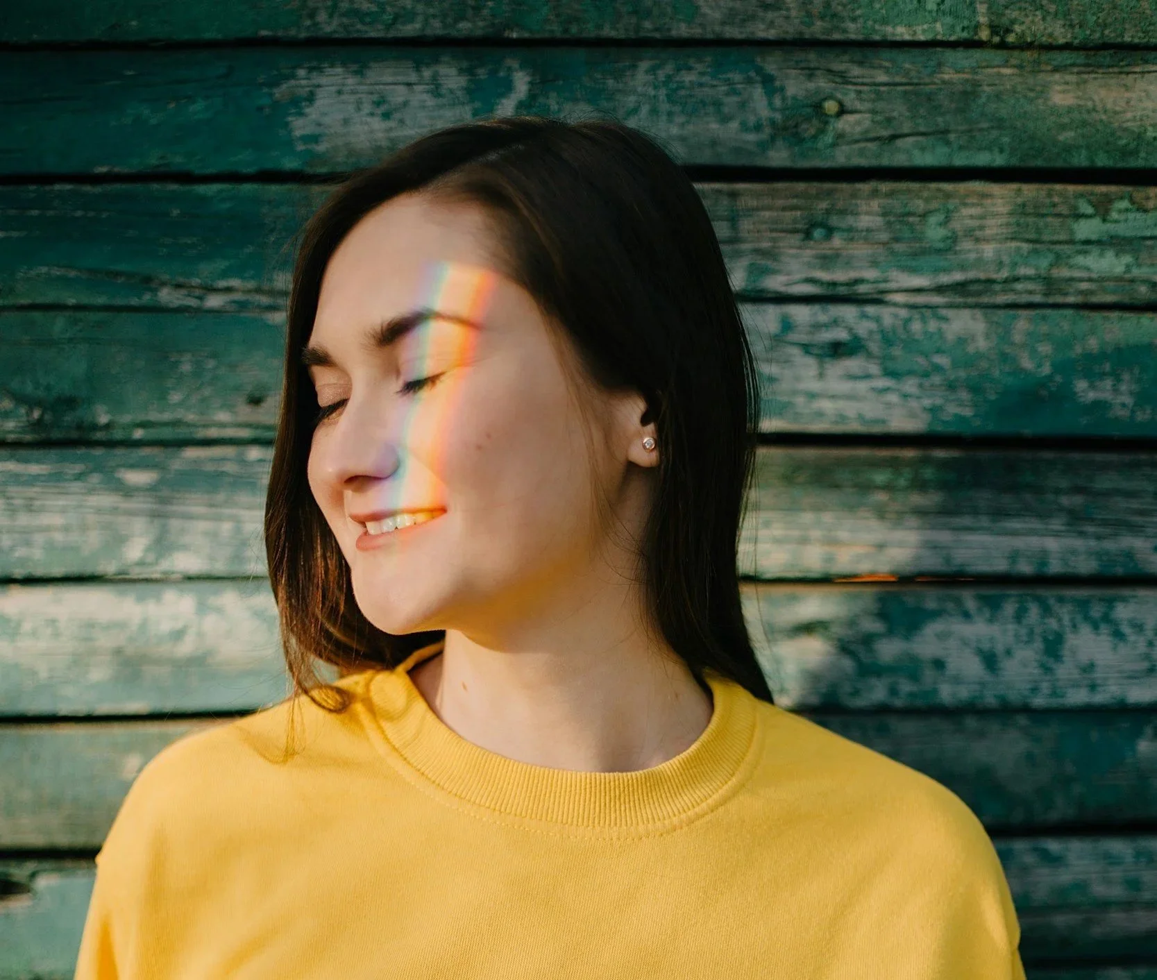 A woman with dark hair, wearing a yellow shirt, smiling with her eyes closed, standing against a weathered wooden background, with a rainbow light reflection across her face.