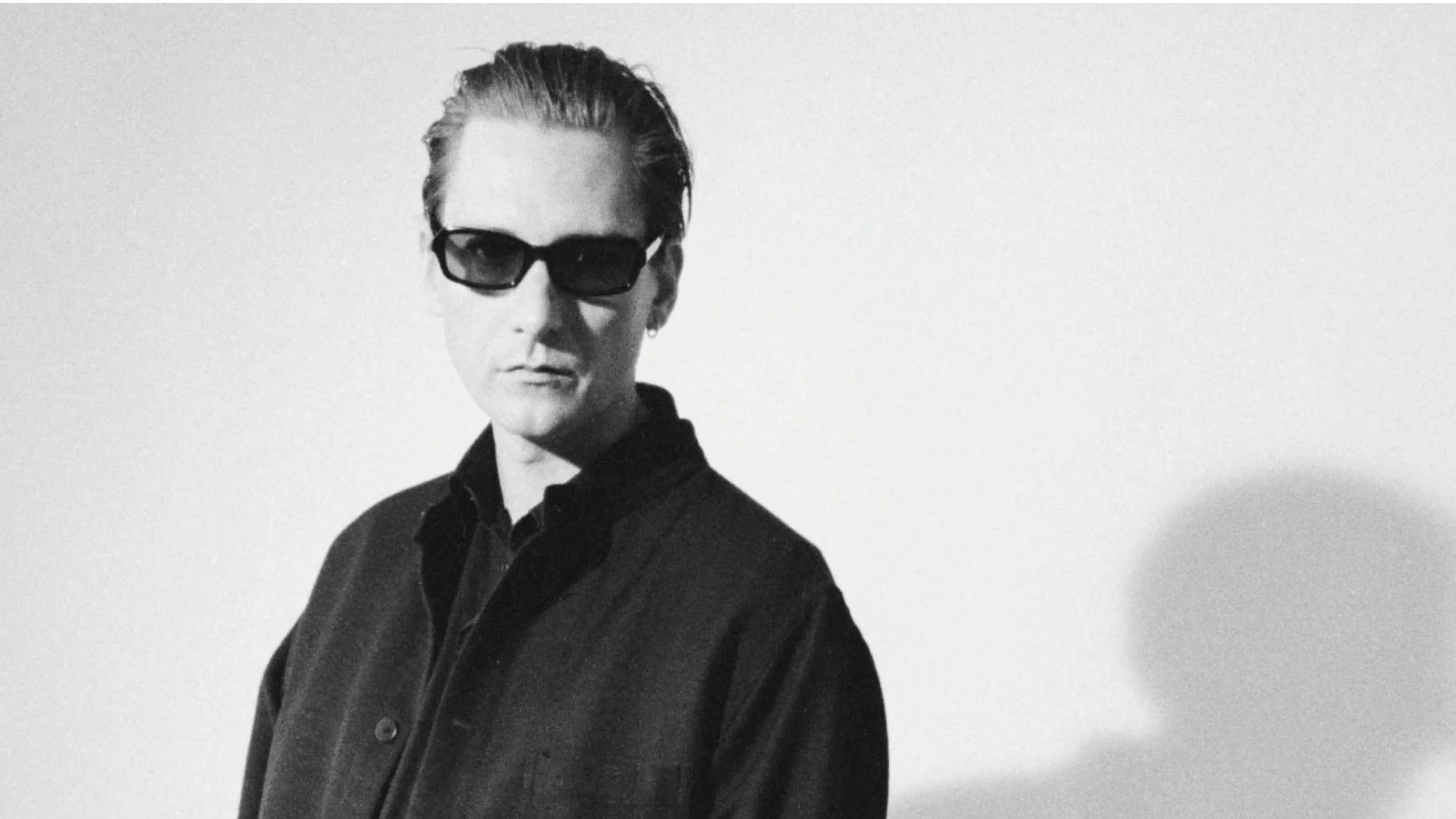 Black and white photo of a woman with slicked-back hair, wearing dark sunglasses, a dark button-up shirt, and a confident expression, standing against a plain light background.