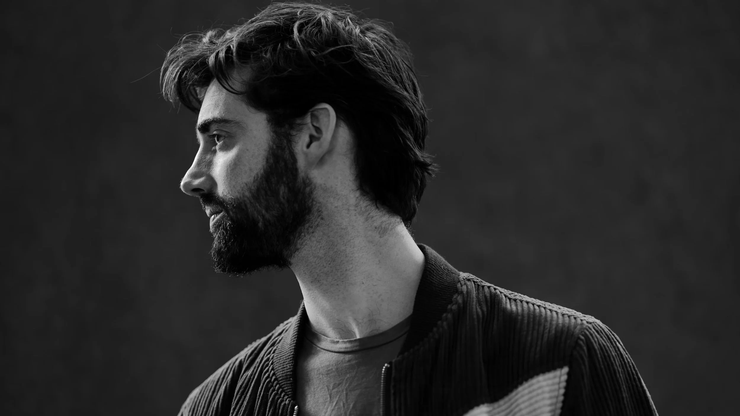 Black and white profile photo of a man with a beard and medium-length hair, wearing a dark jacket and a t-shirt, against a plain dark background.
