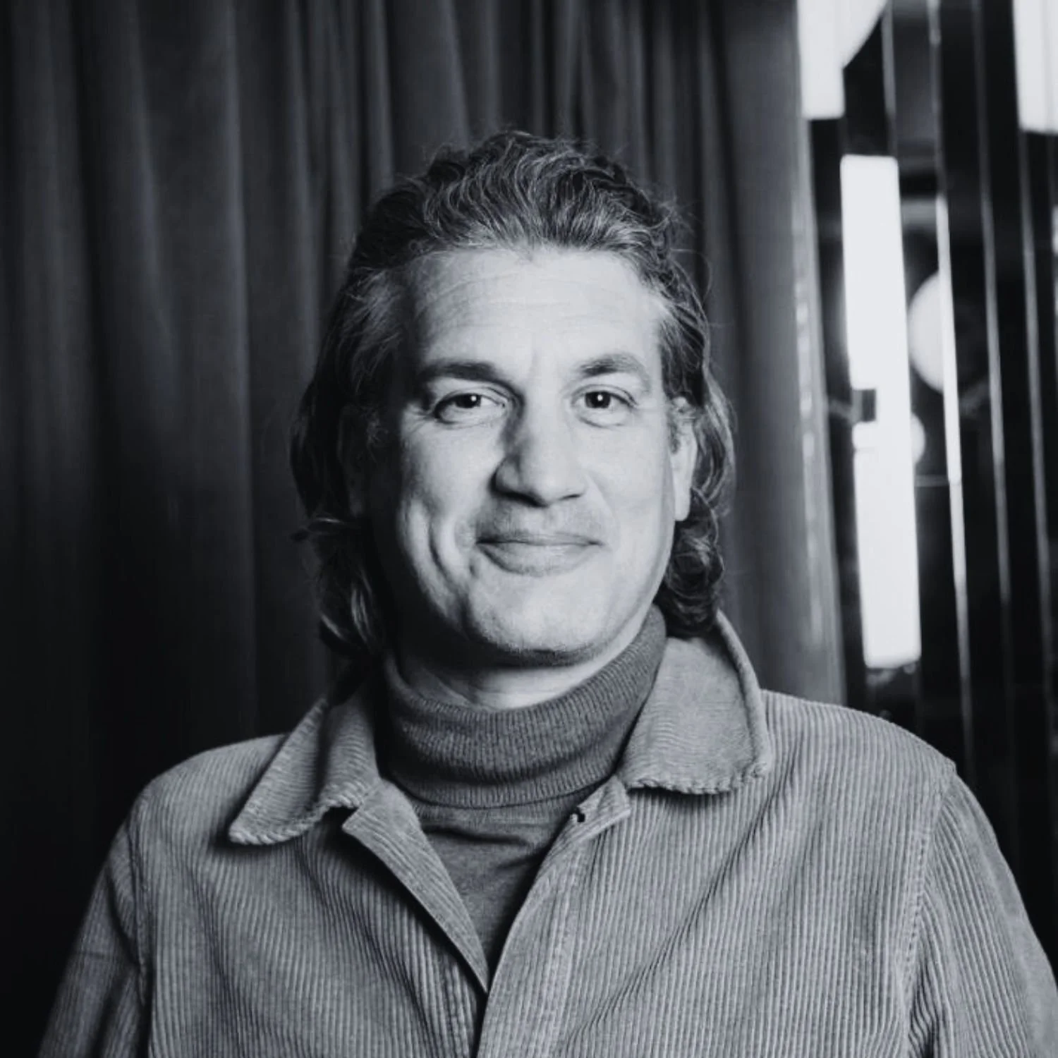 Black and white portrait of a man with shoulder-length hair, wearing a turtleneck and a button-up shirt, standing in front of a wooden background with light coming through a window.