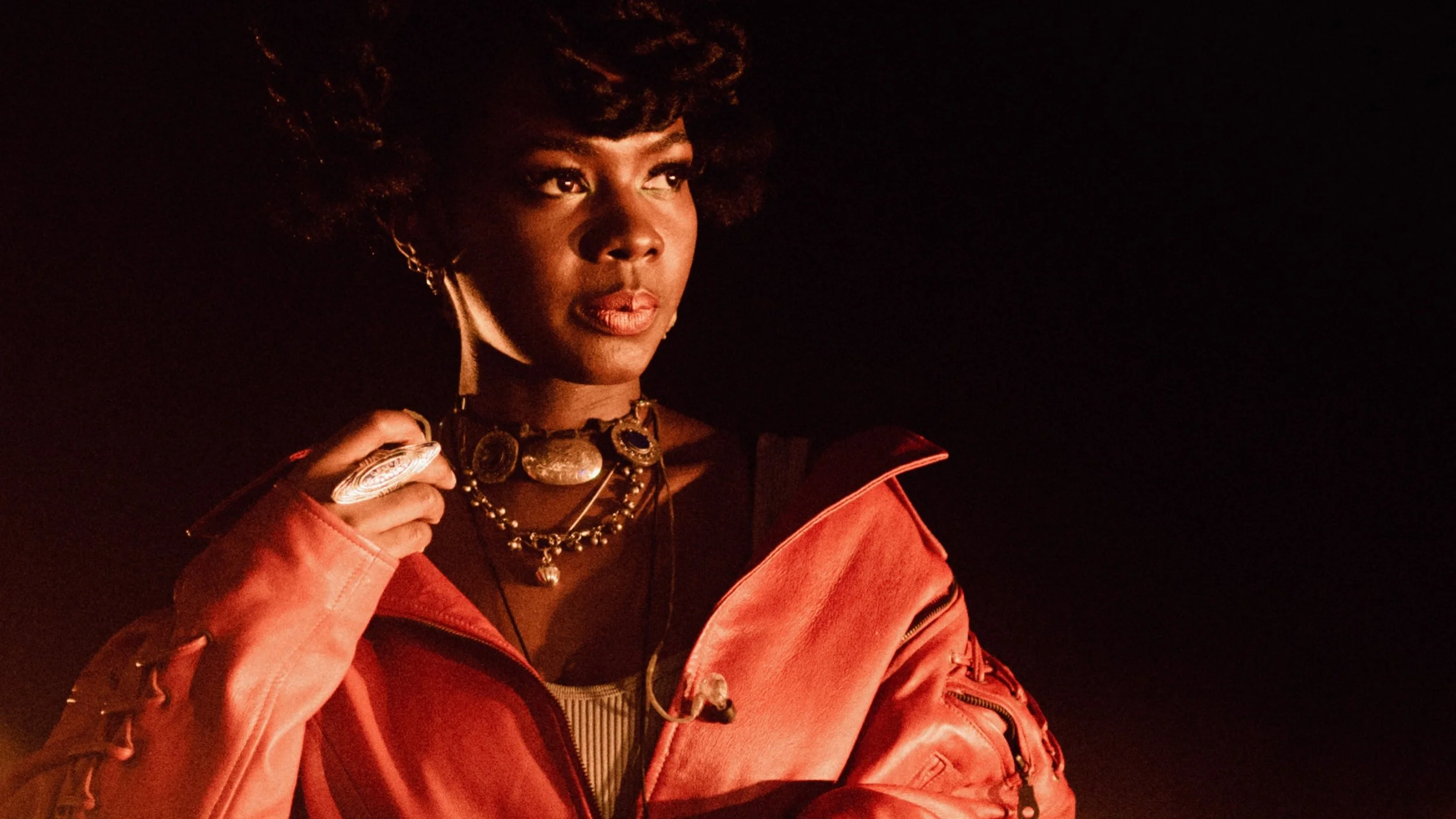 A woman with dark skin and curly hair holding a pocket watch, wearing layered necklaces and a coral pink jacket, against a dark background.