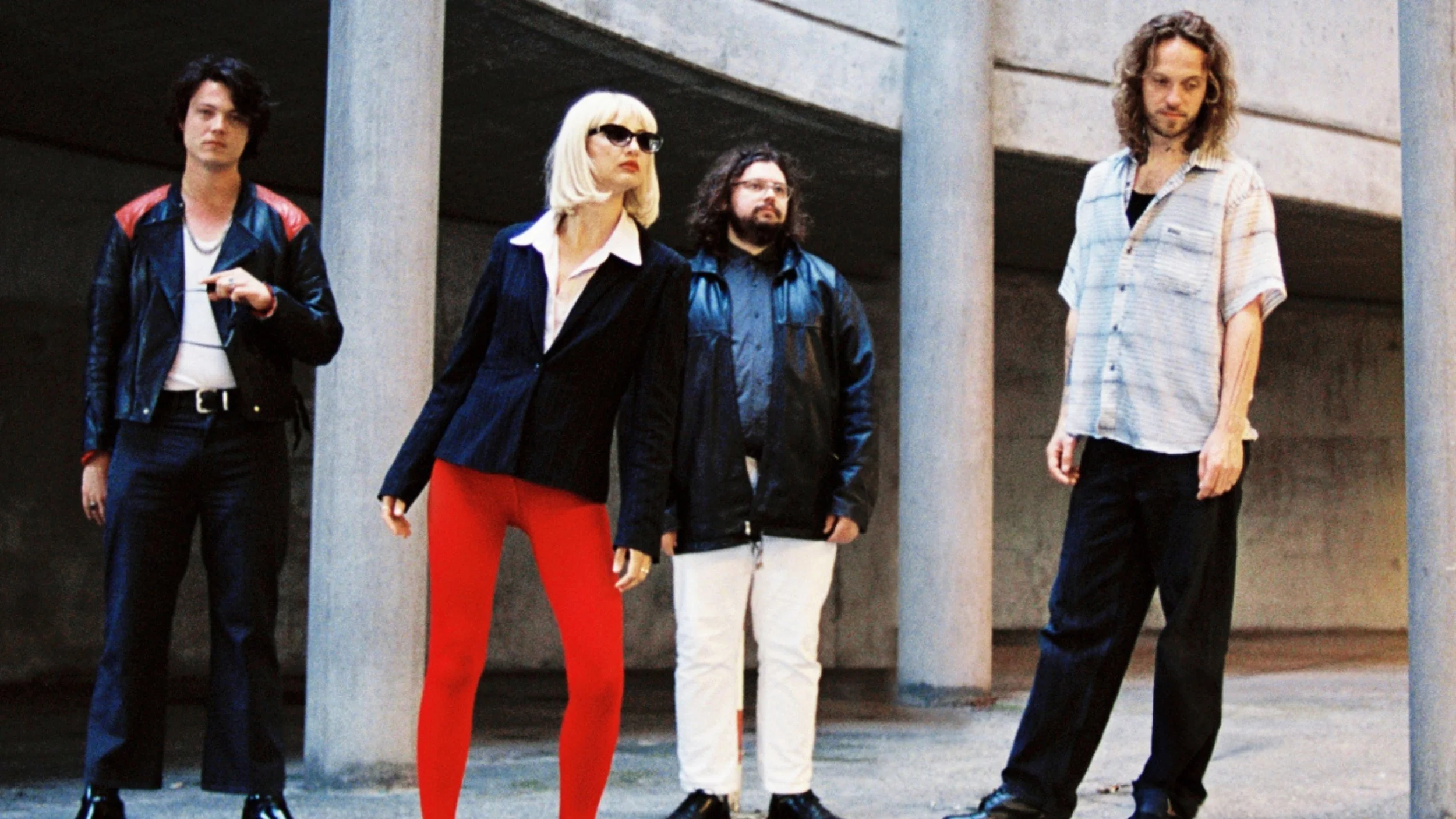 Four people standing outdoors in front of a concrete structure with columns, dressed in 1980s casual and punk fashion.