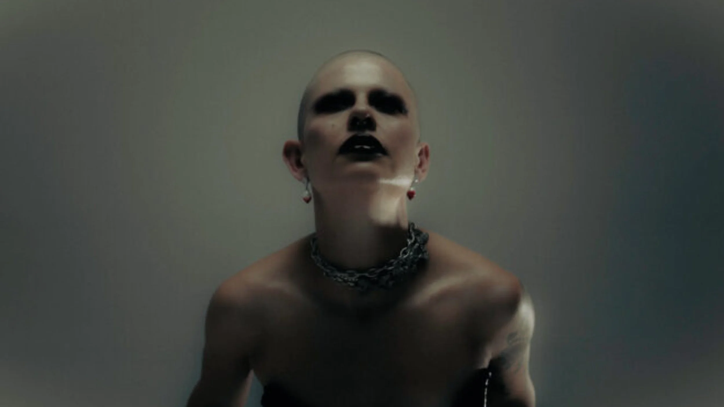 A person with a shaved head and dark makeup on face, wearing black lipstick, earrings, and layered necklaces, posing against a plain background.