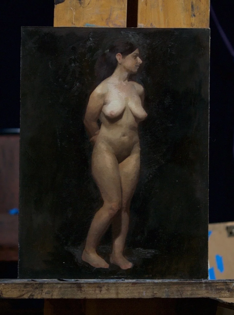 A realistic oil painting of a nude woman standing, facing right, with dark hair, against a black background.