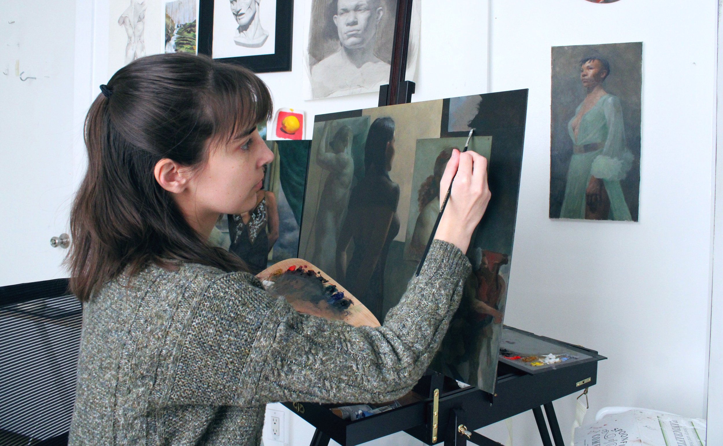 Julia Albo painting realistic portraits of women on canvas in an art studio with portraits and sketches hanging on the wall.