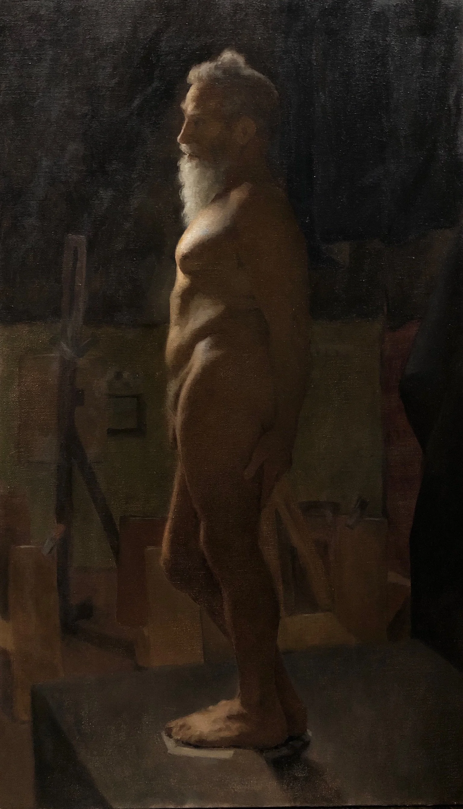 A nude man with white hair and a beard facing left in an artist's studio with art supplies and sketches in the background.