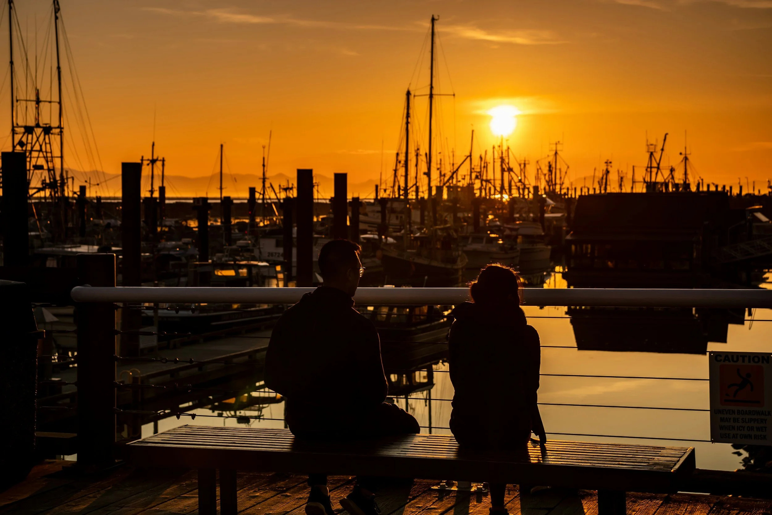Two individuals sitting on a wooden bench on Steveston Village boardwalk against boats during sunset in Richmond