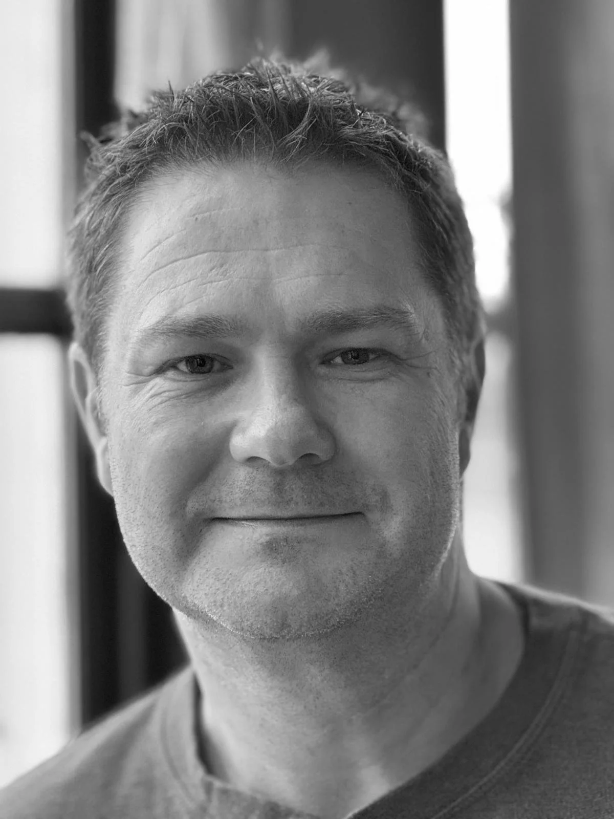 Black and white close-up portrait of a smiling man with short hair, wearing a casual shirt, in front of a window.