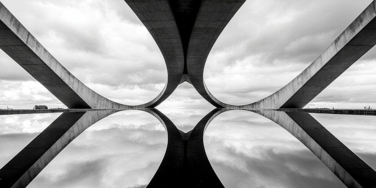 Black and white photo of a modern bridge with curved concrete supports reflected in a calm water surface.