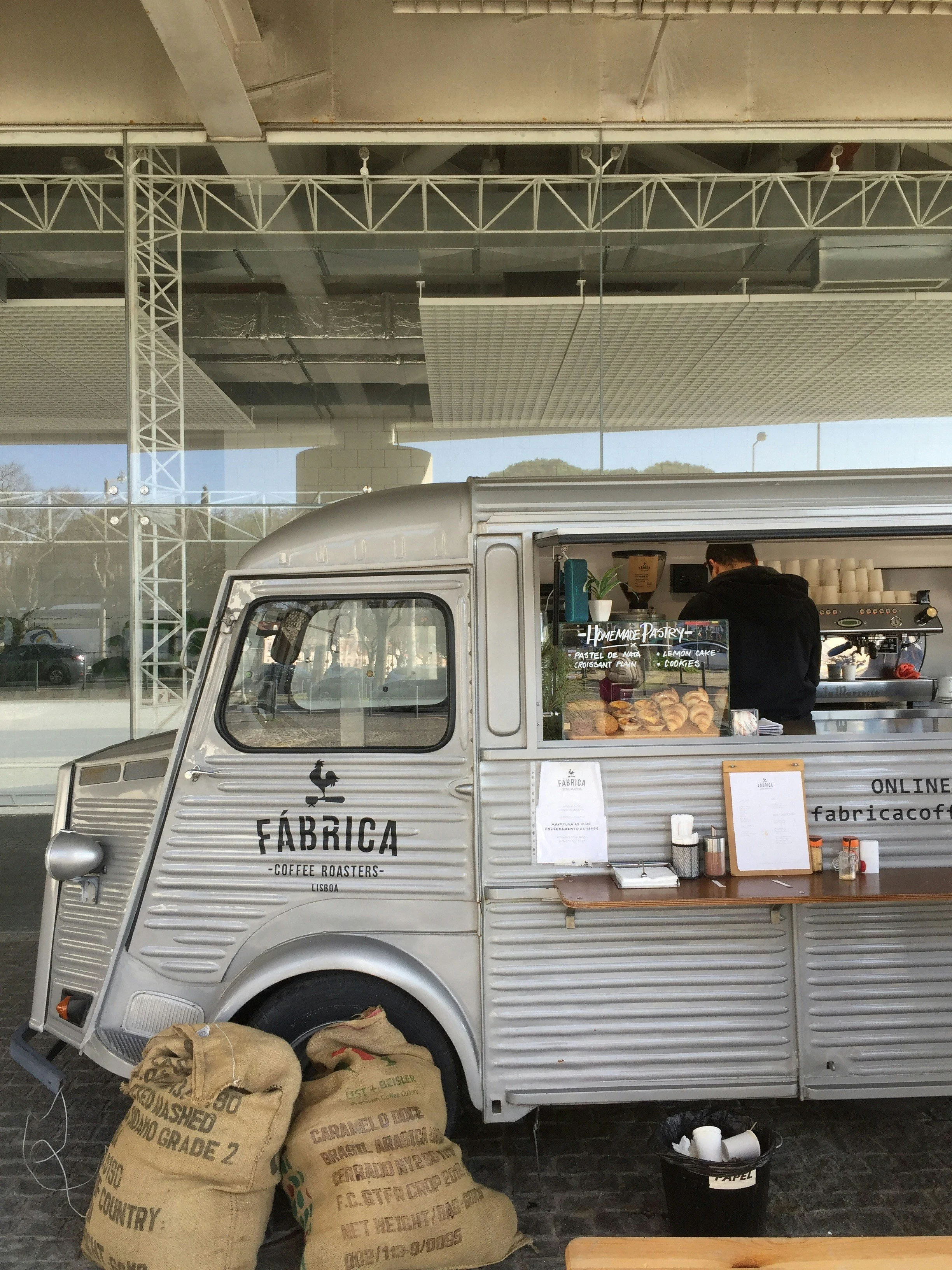 A silver food truck parked outside a building with a large window. The truck is serving handmade pastries, including croissants, lemon cake, and cookies. There are sacks of coffee beans near the front wheel and a small black bucket underneath. Inside the truck, a barista is preparing coffee.