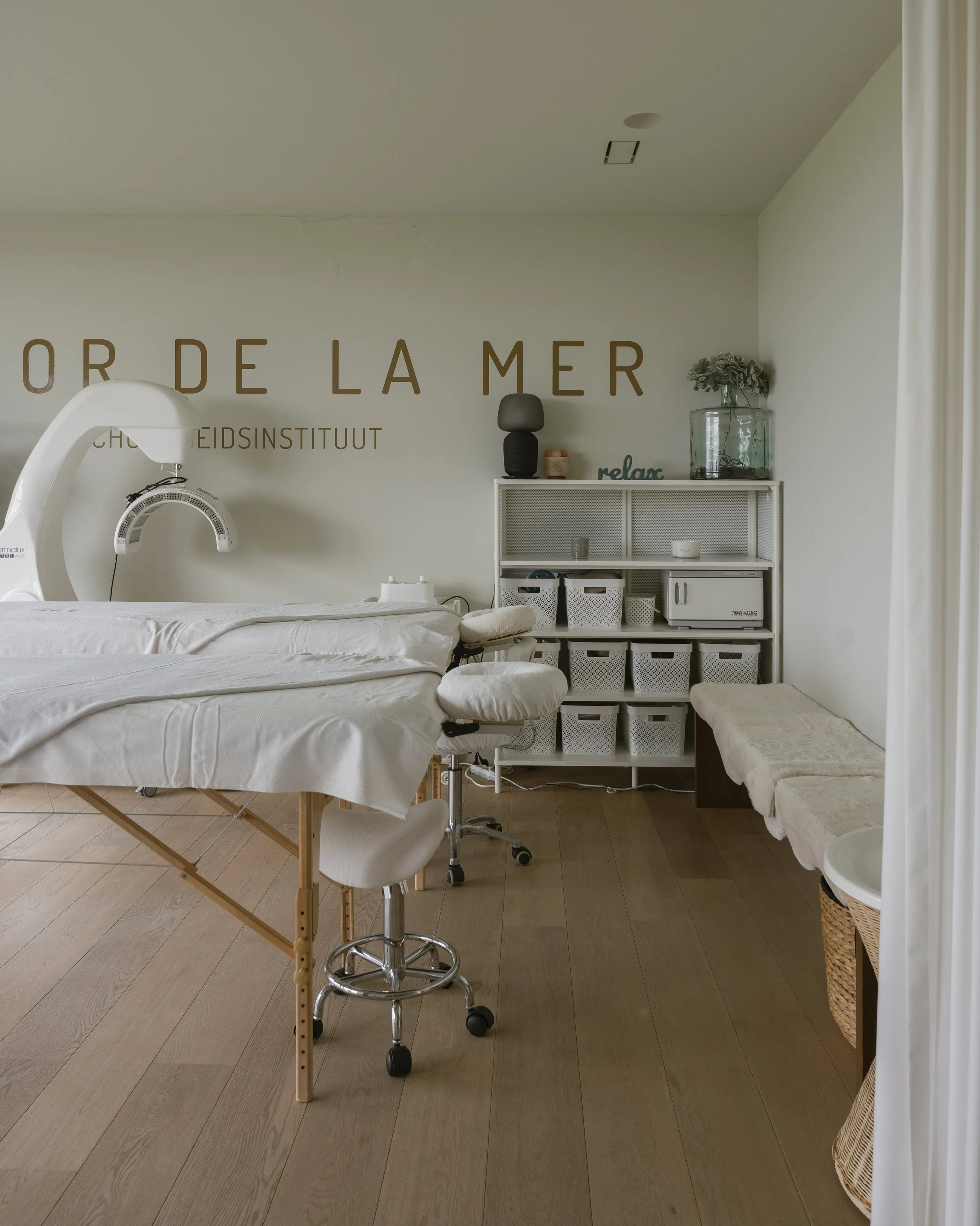 Een rustgevende behandelkamer met een massage- of behandeltafel, een bureaustoel, opbergruimte met manden, een bank en decoratieve planten. Muurtekst leest 'ordre de la mer' en 'relax'.