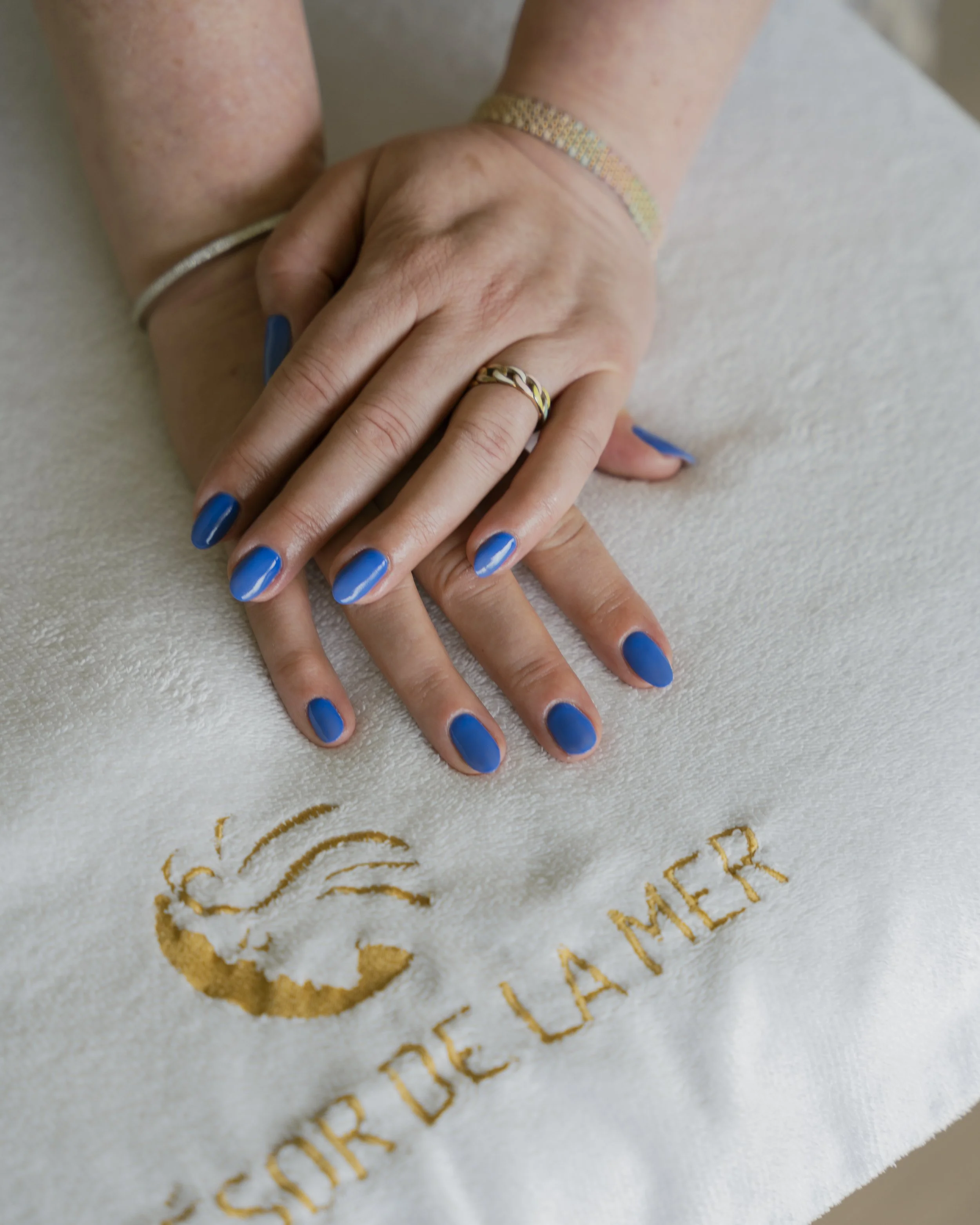 Twee handen met blauwe nagellak, sieraden en een logo met de tekst 'SORDELA MER' op een witte handdoek.