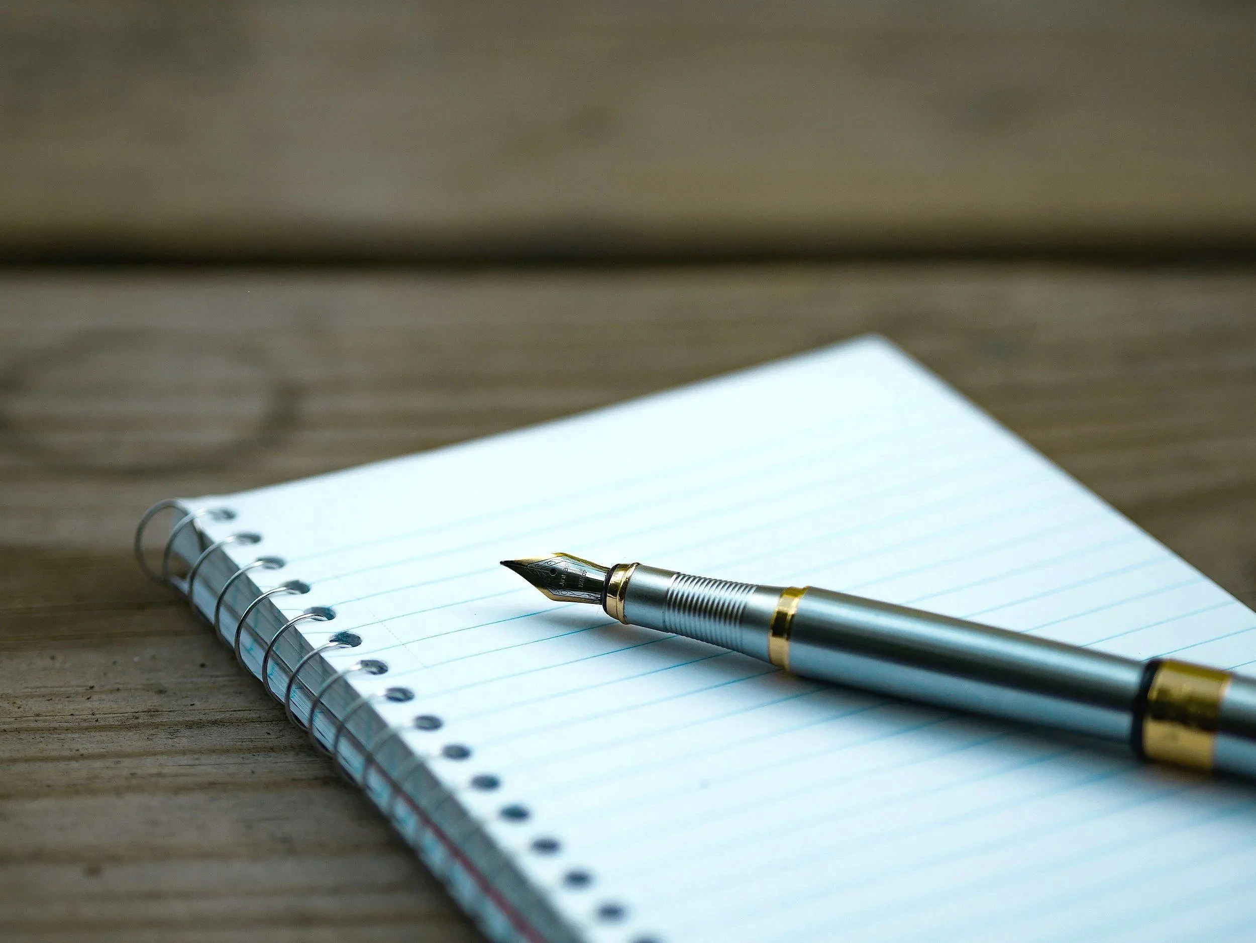 A silver fountain pen resting on a lined spiral notebook on a wooden surface.