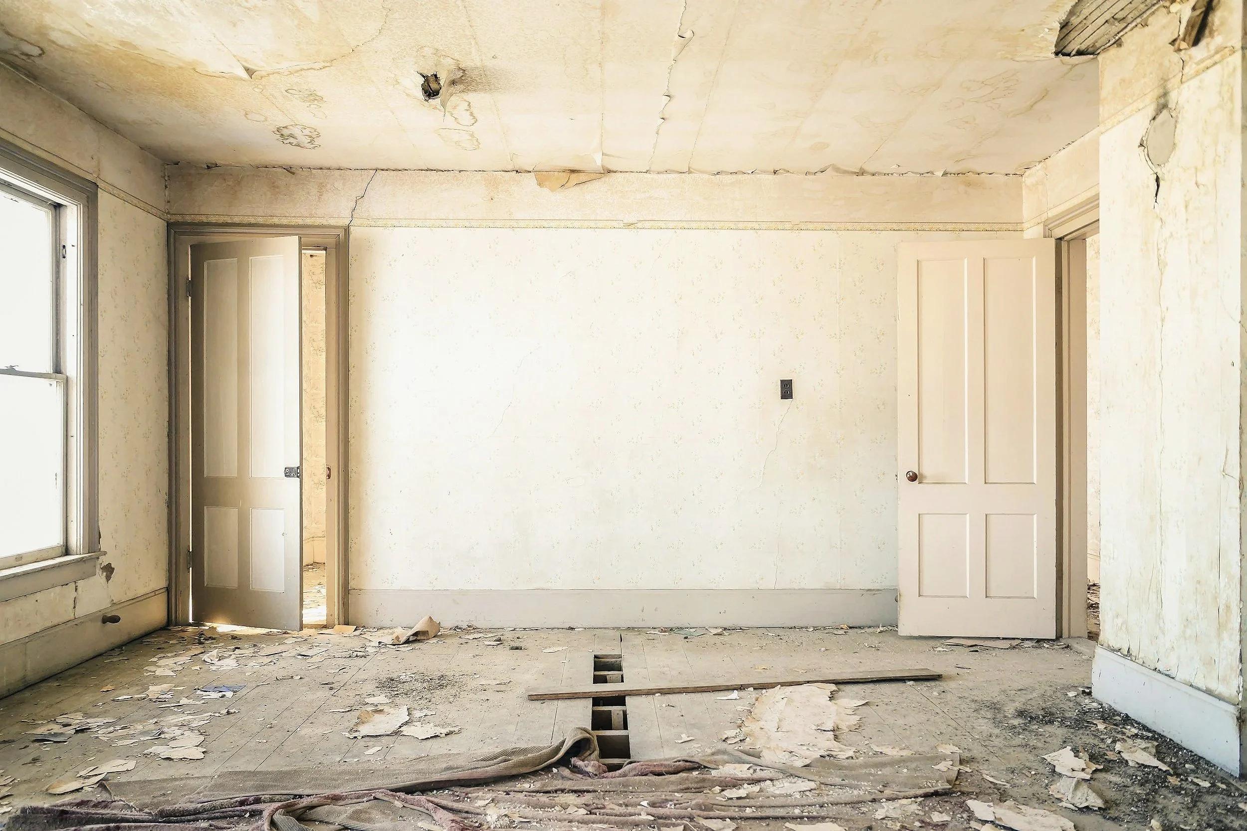 Empty room in a dilapidated house with peeling wallpaper, broken flooring, and open doorways.