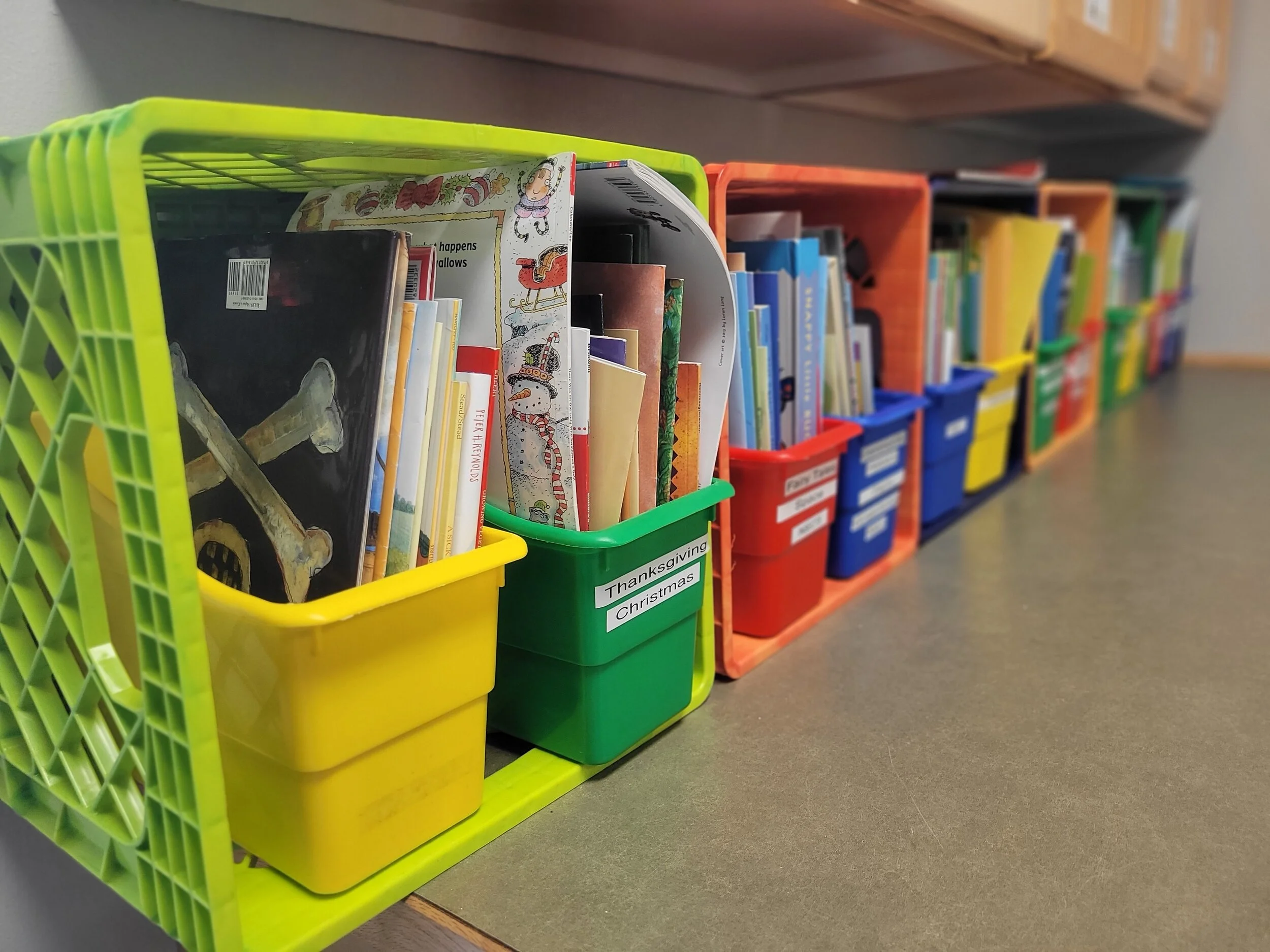 Colorful plastic bins filled with books, placed on a gray surface in a library or classroom setting.