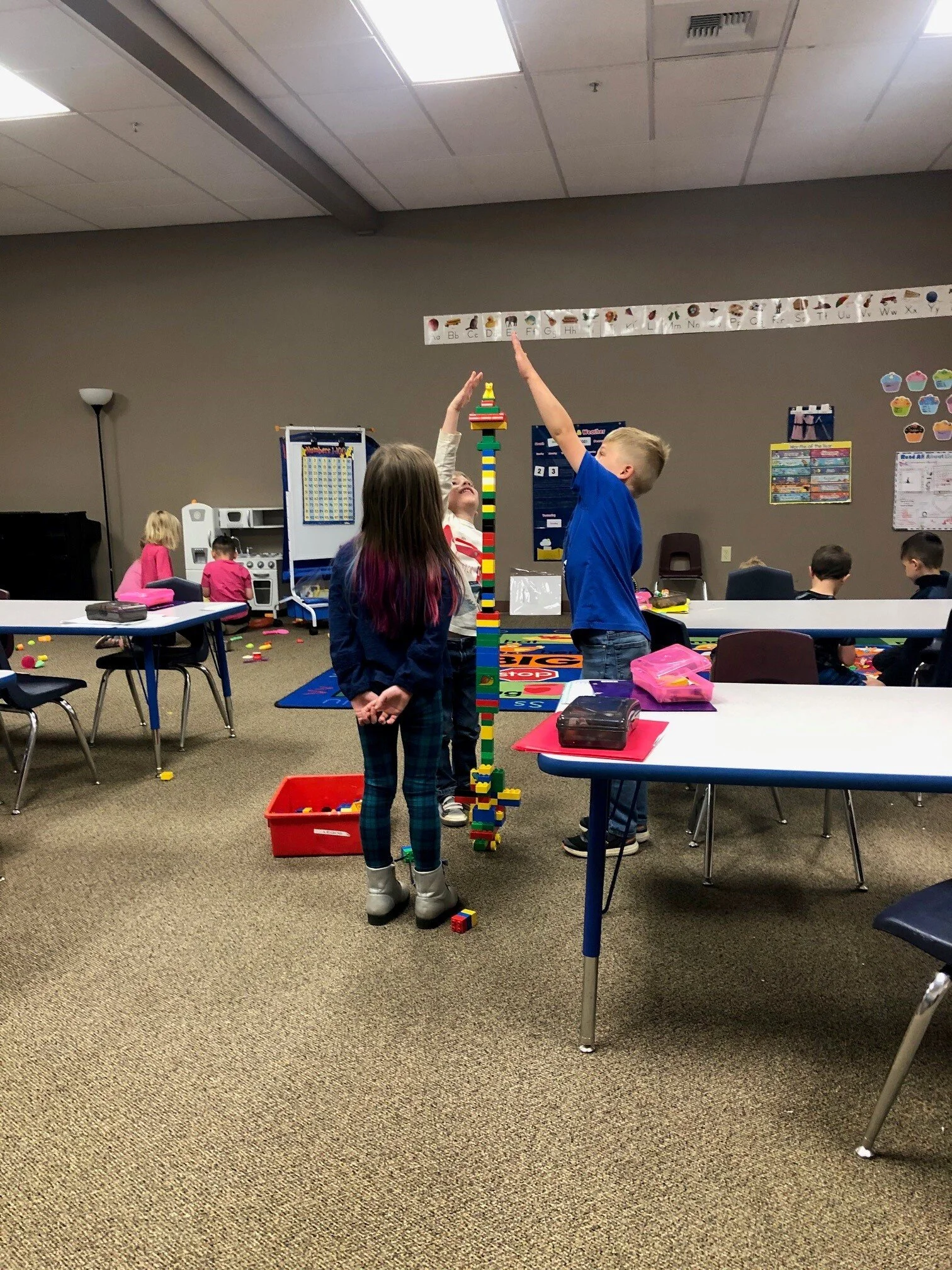 Three children building a tall tower with colorful LEGO bricks in a classroom.