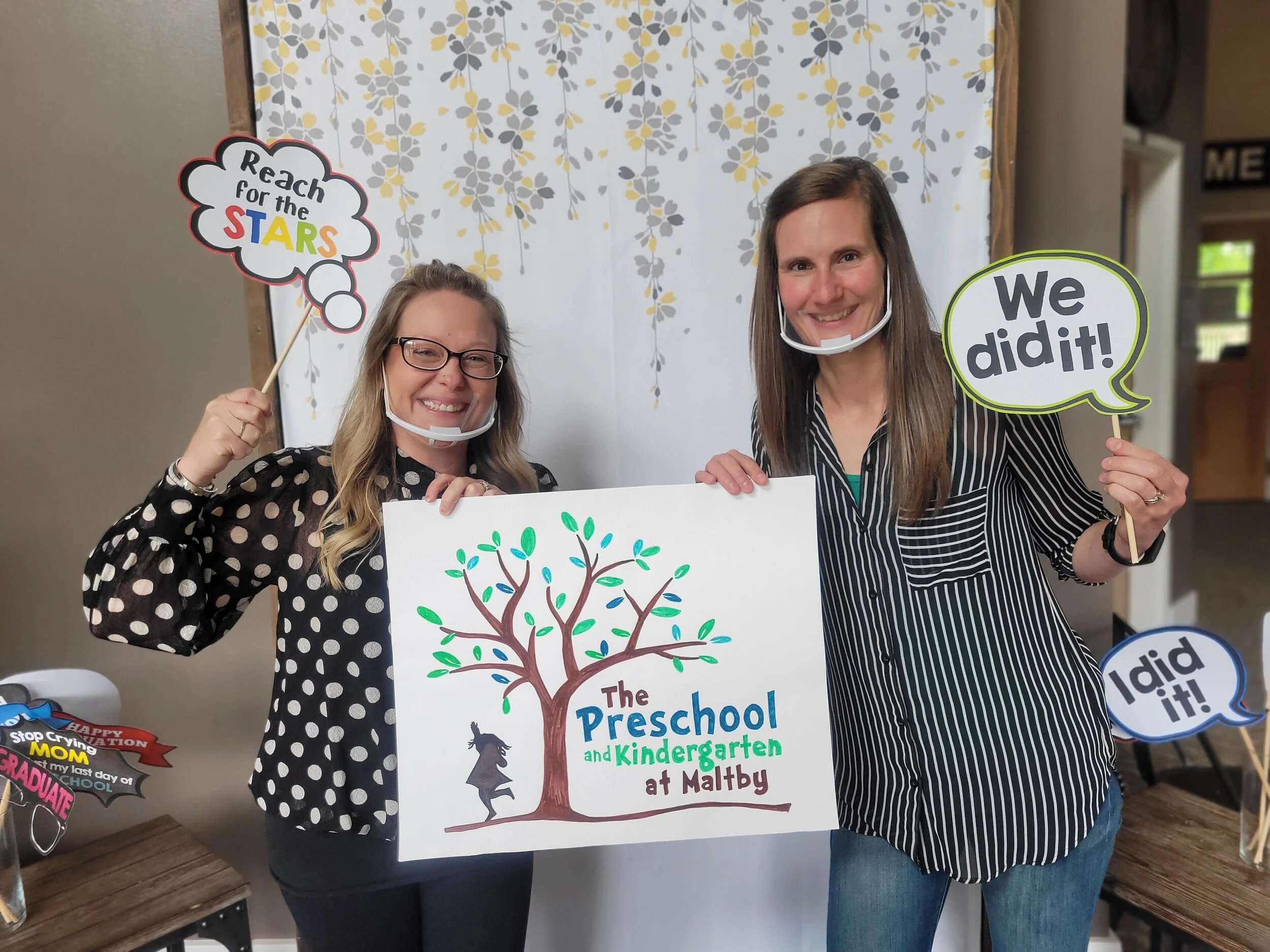 Two women standing in front of a white tree mural, holding signs that say "Reach for the STARS" and "We did it!", celebrating a preschool and kindergarten graduation at Maltby.