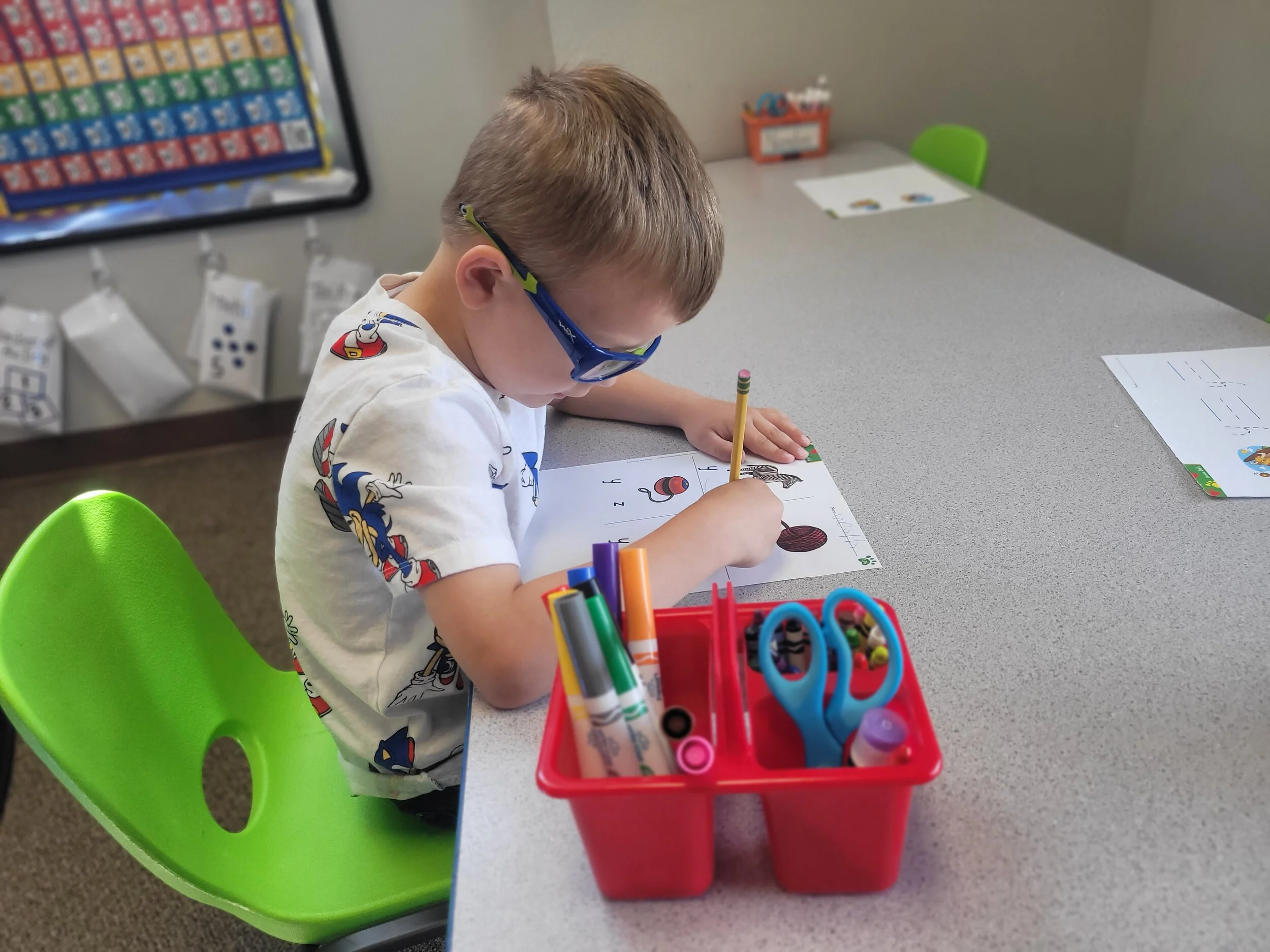 Young boy with glasses coloring at a classroom table with school supplies and educational posters.
