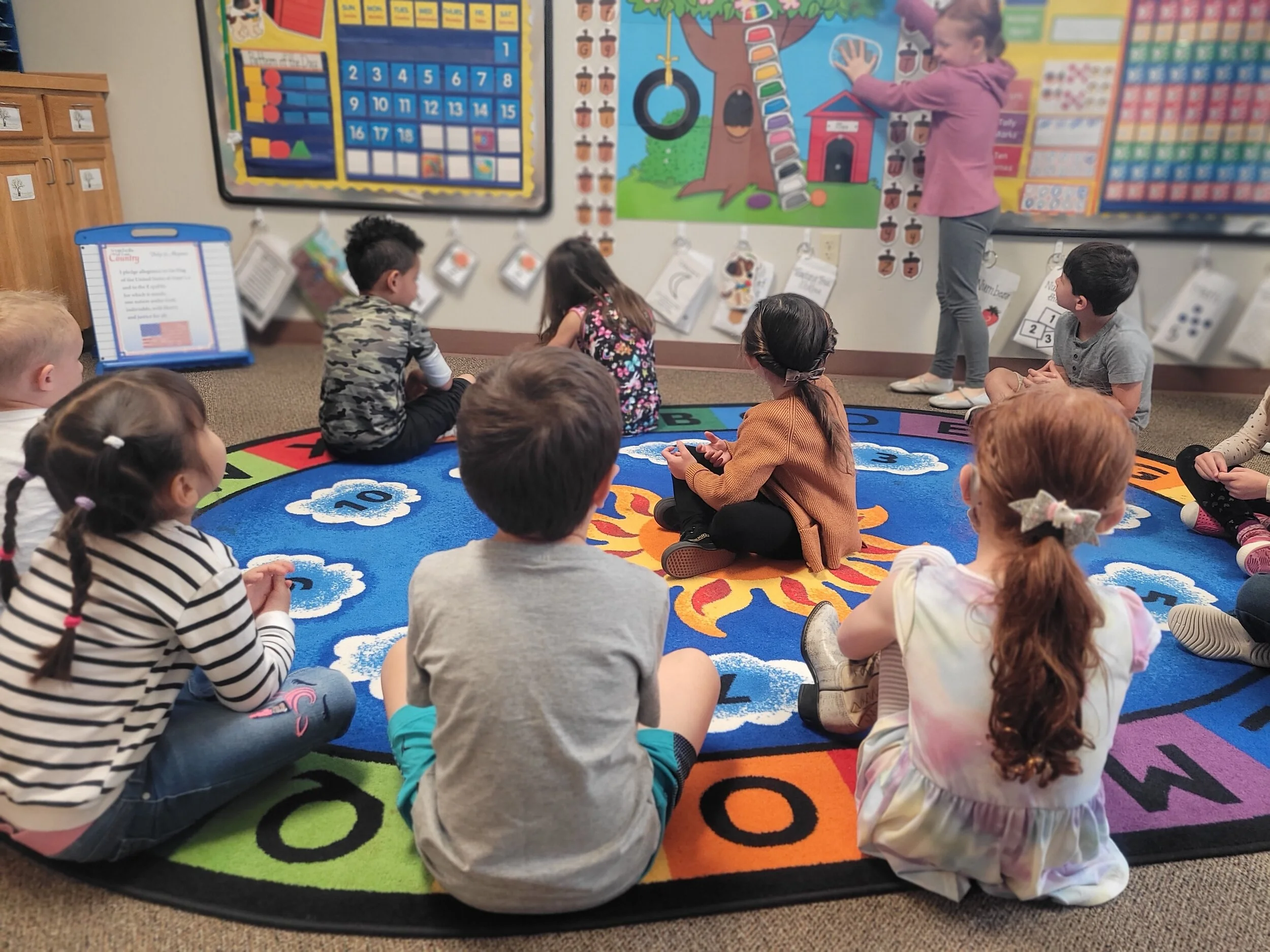 Children sitting cross-legged on a colorful rug in a classroom, watching a girl in a pink sweater presenting near a wall with educational posters.