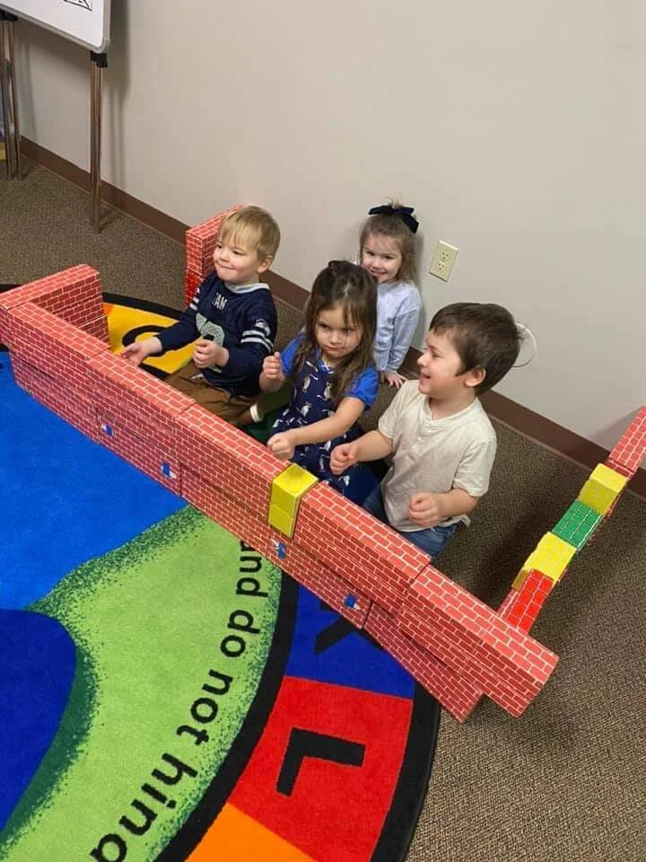 Four young children sitting behind foam blocks designed to resemble a brick wall, on a colorful circular rug in an indoor setting.