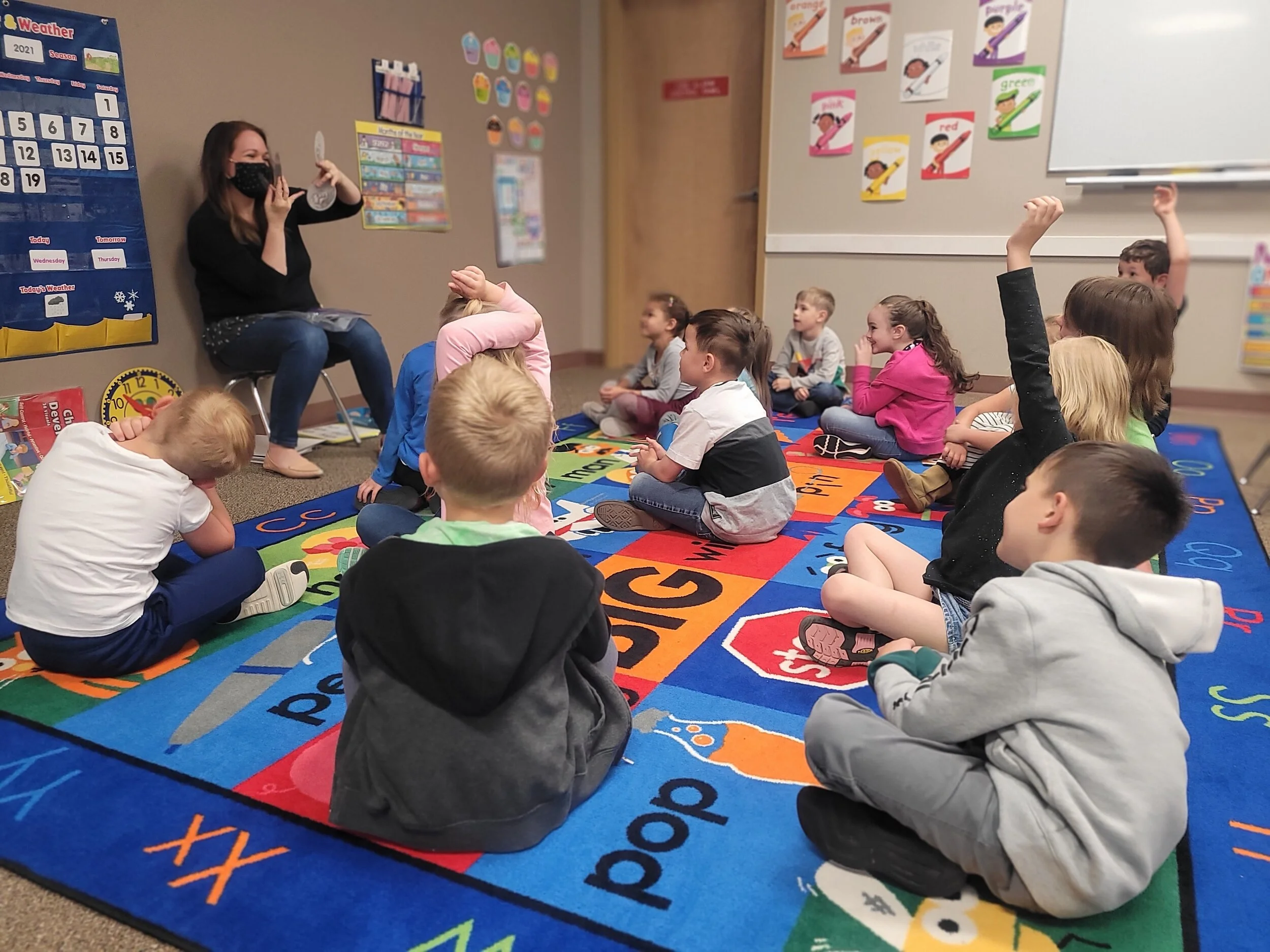 A teacher reading a book to a group of young students seated on a colorful alphabet-themed rug in a classroom, with some children raising their hands.