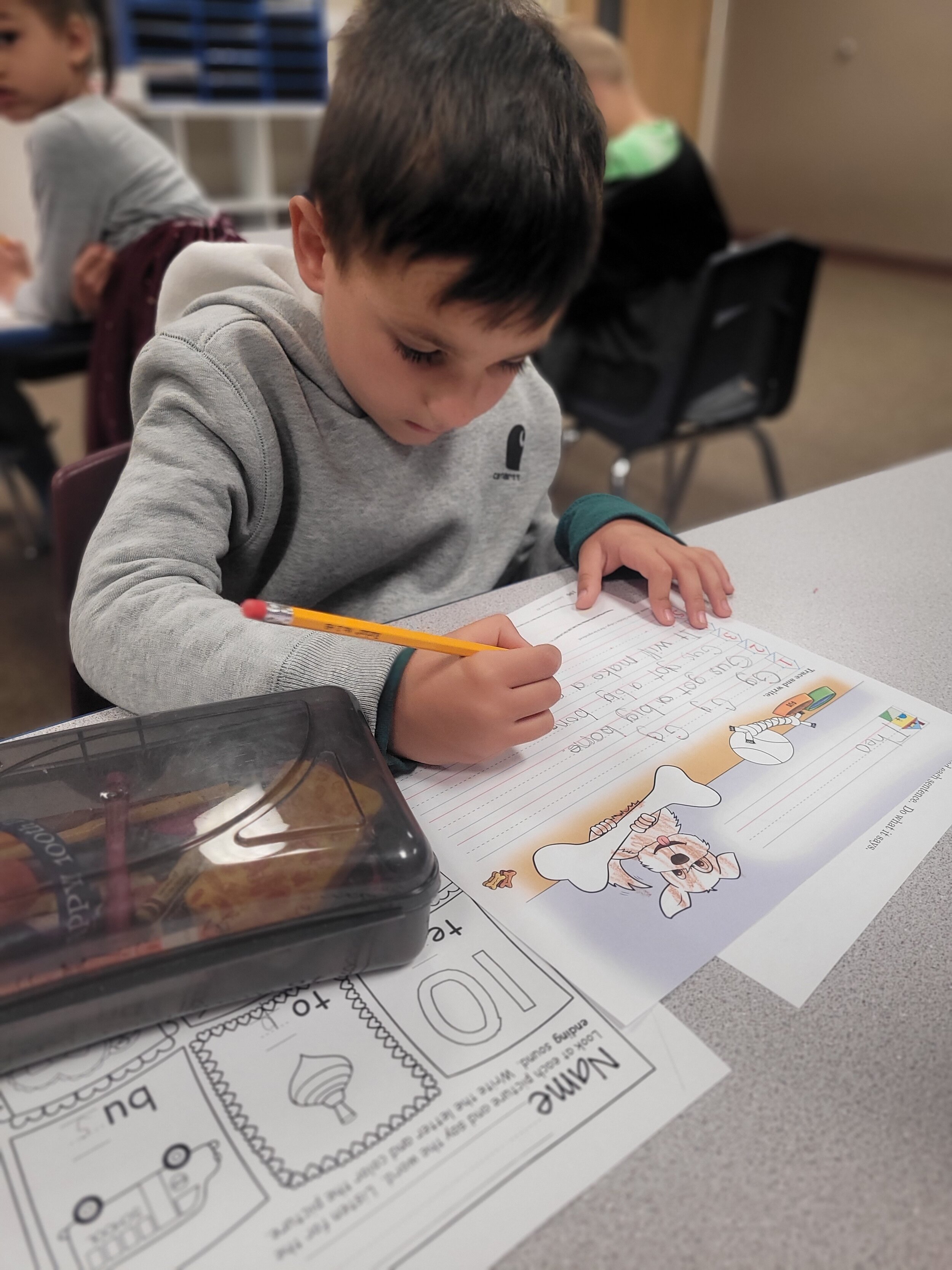 A young boy in a gray hoodie writing on a colorful worksheet featuring a cartoon dog at a classroom desk. Other children are visible in the background.