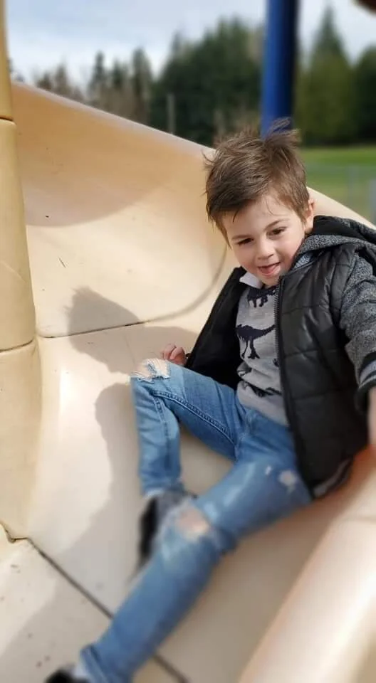 A young boy with brown hair, wearing a gray sweatshirt decorated with black dinosaur silhouettes, a black puffer jacket, and ripped blue jeans, sitting on a beige playground slide outdoors. He is smiling and leaning back slightly, with a background o