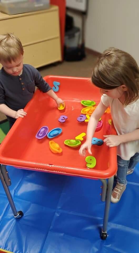 Two children playing with colorful foam numbers on a red plastic table in a classroom.