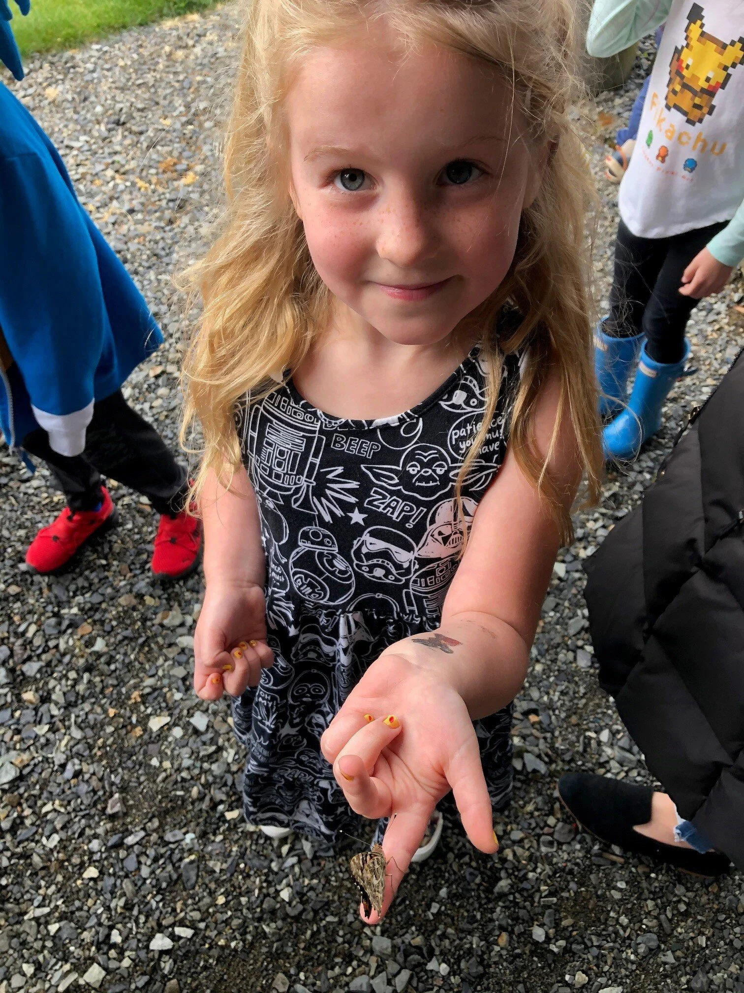 A young girl with long blonde hair and bright blue eyes is holding a moth on her hand. She is smiling and looking at the camera. She is wearing a black sleeveless dress with white graphics and fall-colored nail polish. Other children are visible in t