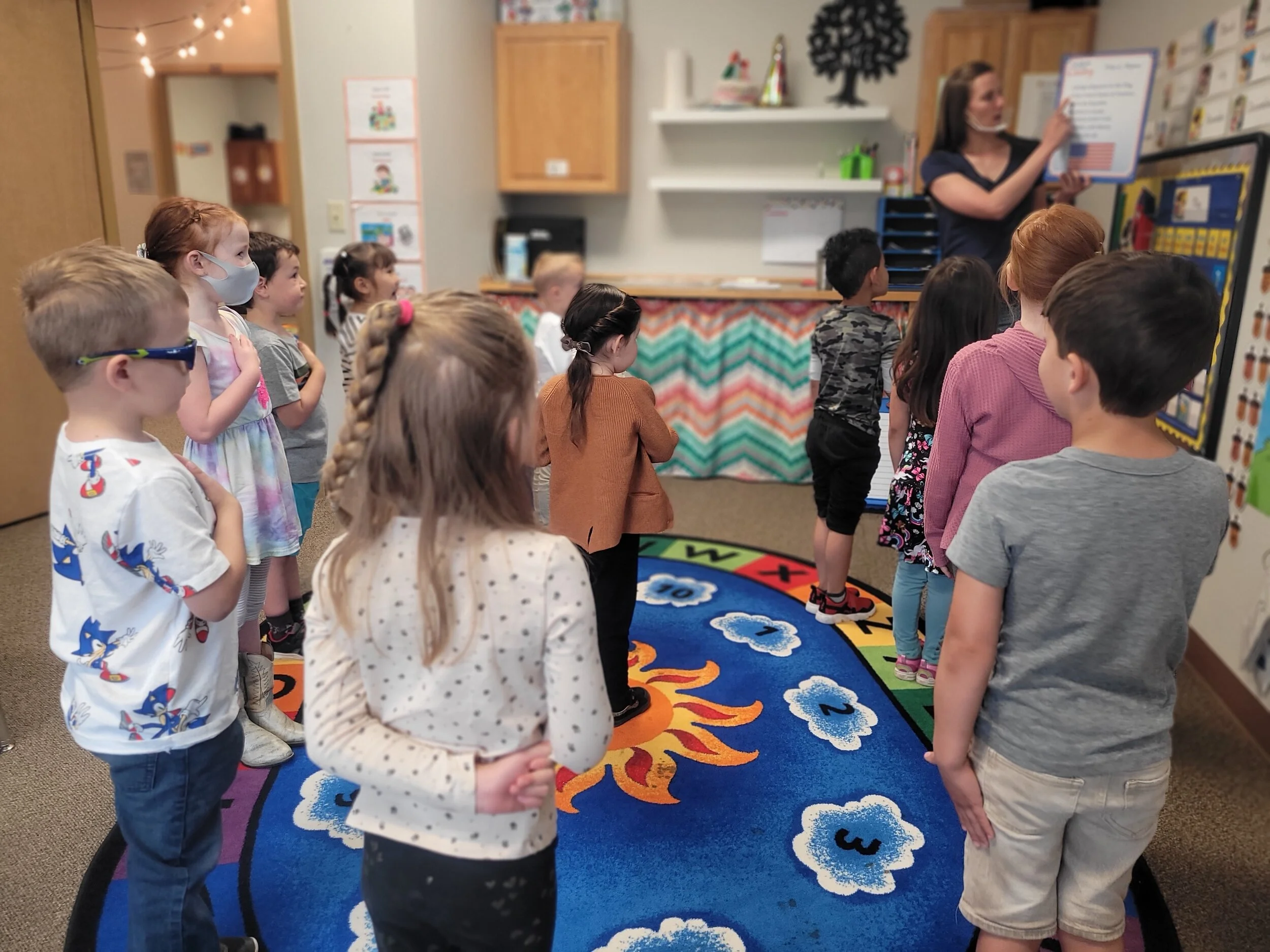Group of children standing on a colorful rug in a classroom, facing a teacher who is reading from a large book, with educational posters and decorations in the background.