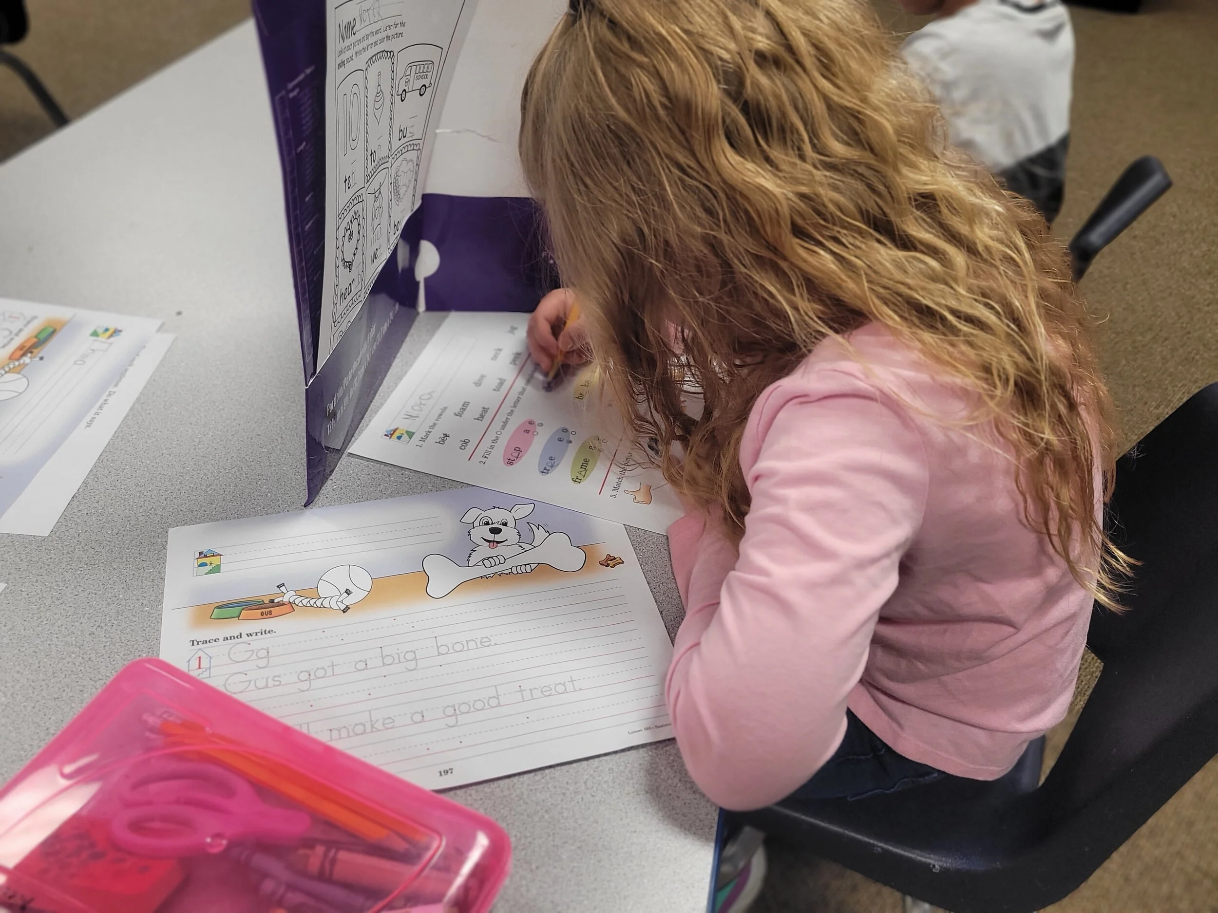 A young girl with curly red hair wearing a pink shirt, sitting at a desk, writing on a worksheet with a cartoon dog and bone illustration. A pink pencil case is on the desk. There are additional worksheets and a display stand with educational content