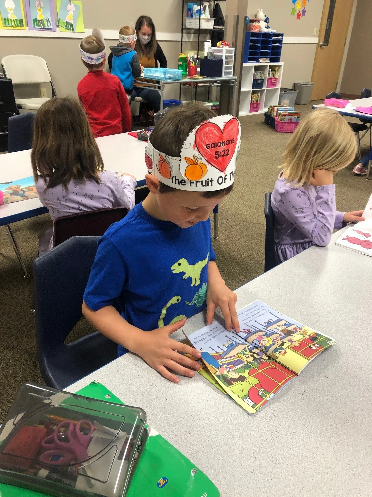 A young boy wearing a paper crown, decorated with pumpkins and gospel message, reads a colorful children's storybook at a classroom table, with other children and a teacher in the background.