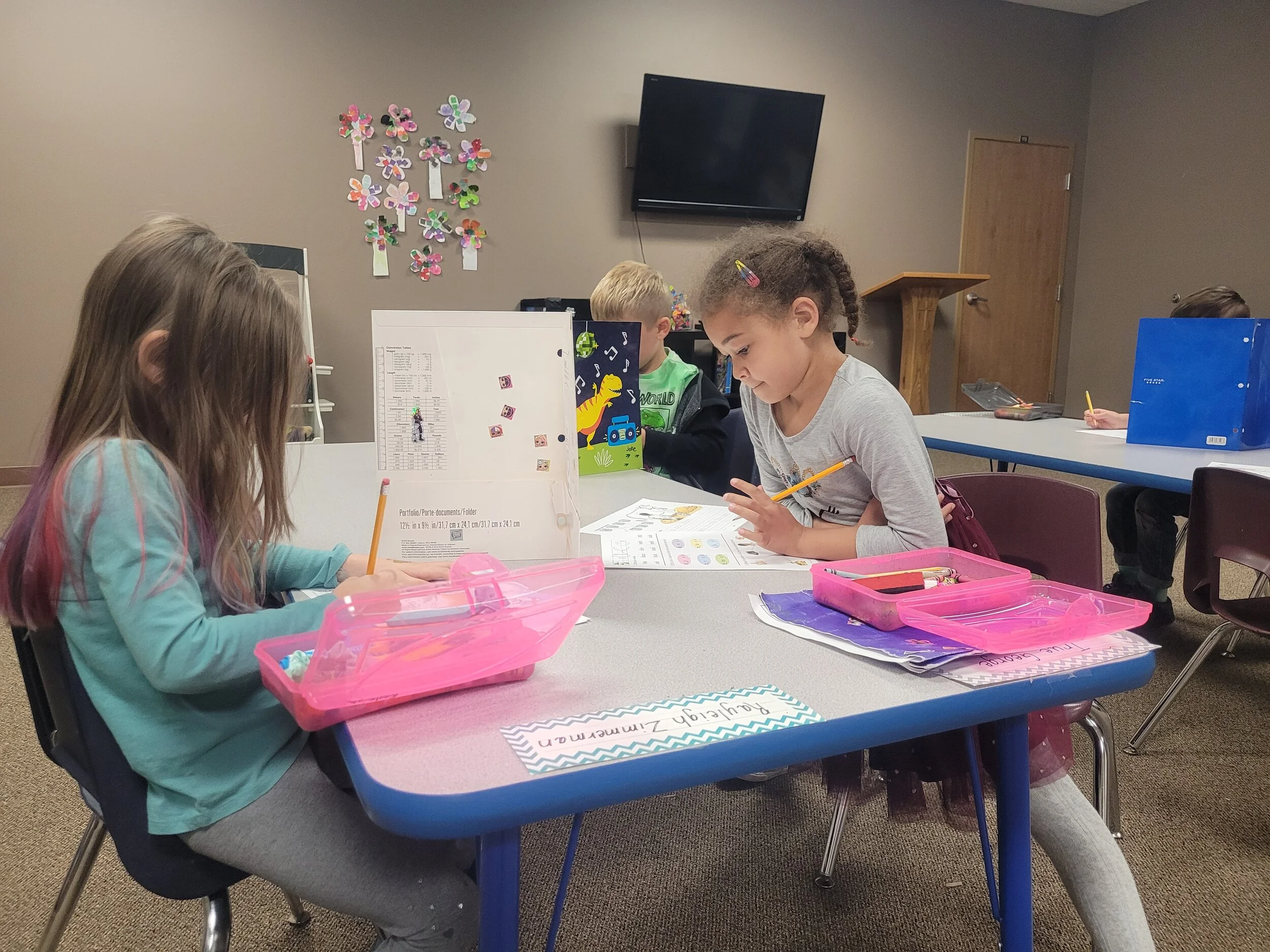 Four children sitting at a table, working on assignments with school supplies and pink pencil cases. A classroom with wall decorations, a TV, a door, and a podium in the background.