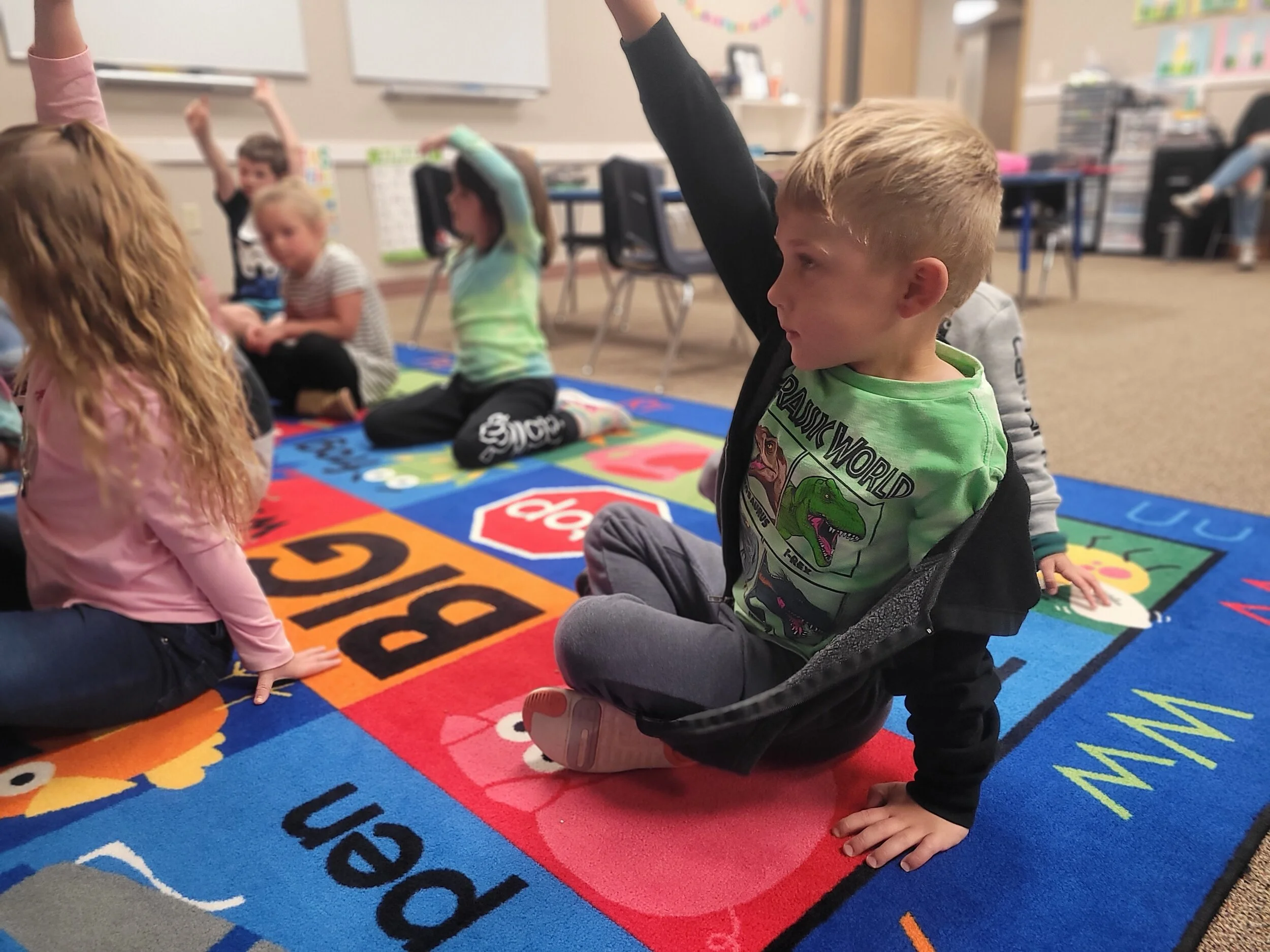 Children sitting on a colorful alphabet-themed rug in a classroom, raising their hands during a lesson.