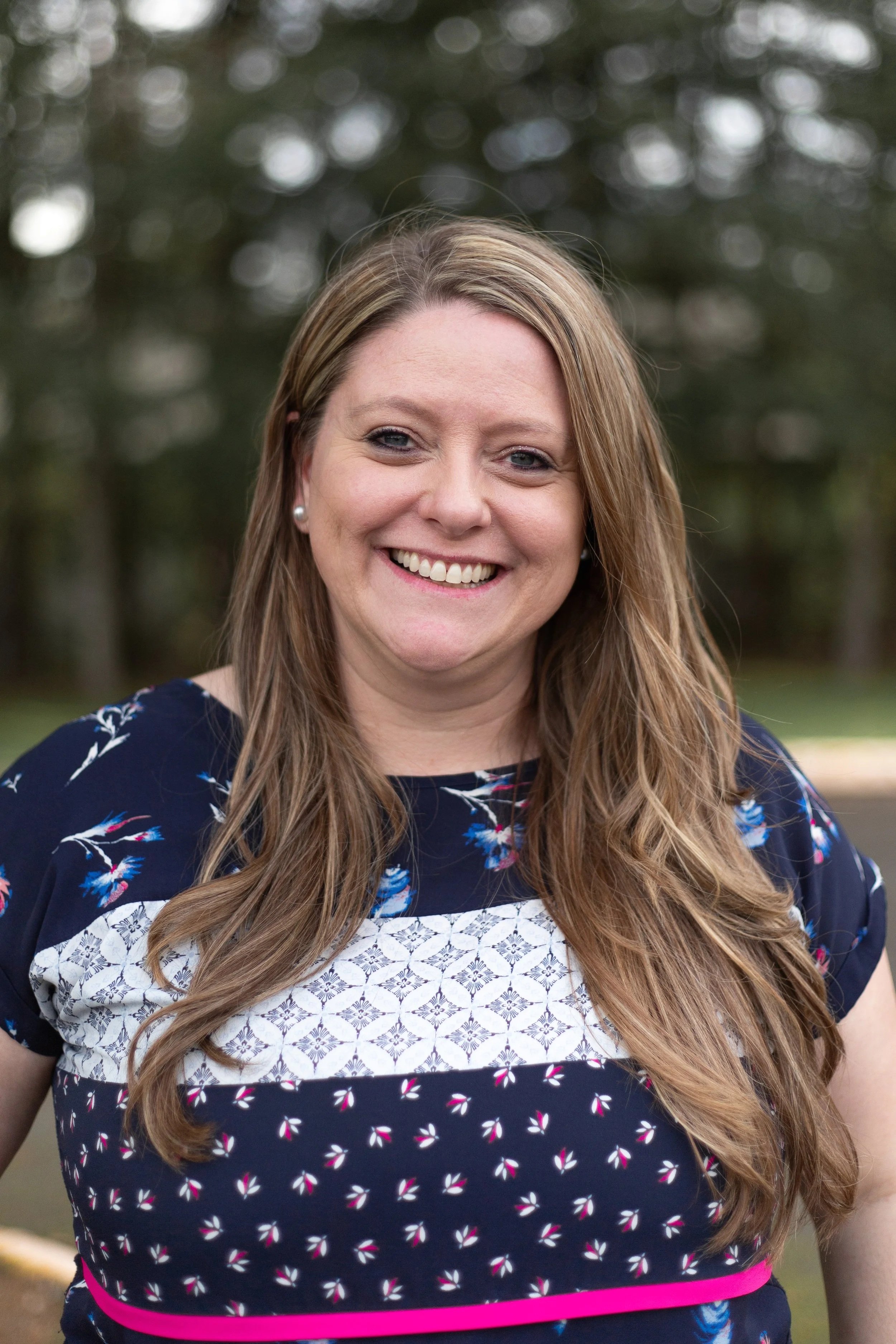 A smiling woman with long, wavy brown hair, wearing a navy blue dress with pink, white, and blue patterns, standing outdoors with blurred trees in the background.