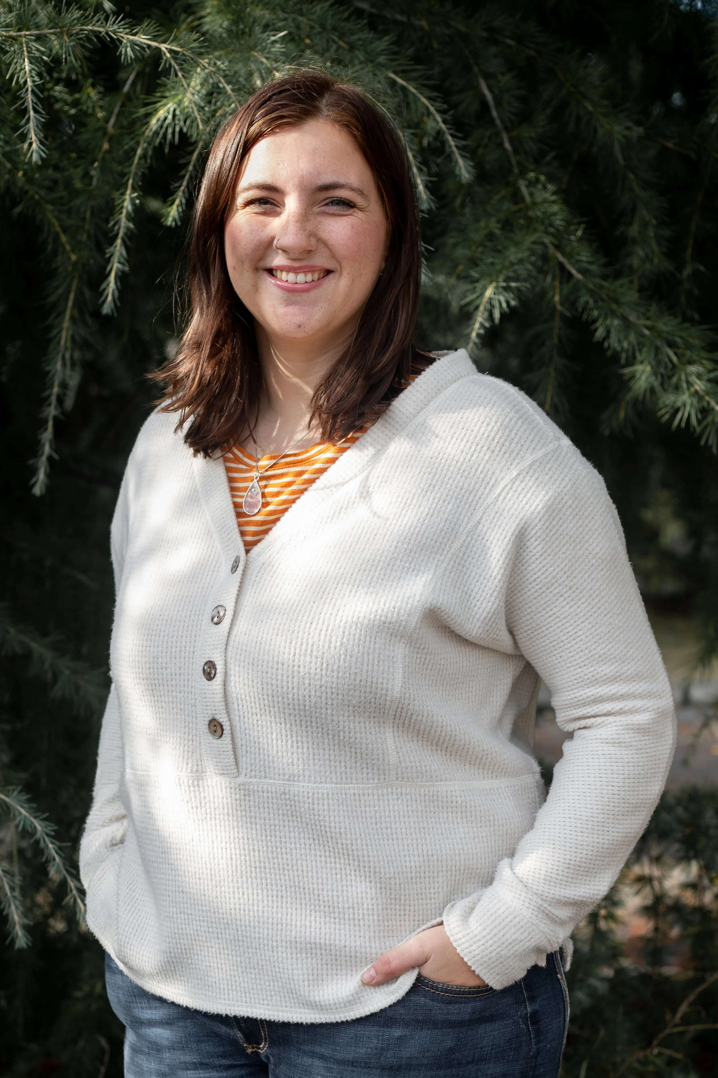 A young woman with shoulder-length brown hair and light skin, smiling and standing outdoors in front of green pine tree branches. She is wearing a white, long-sleeved, button-up sweater over a striped orange and white shirt and jeans.