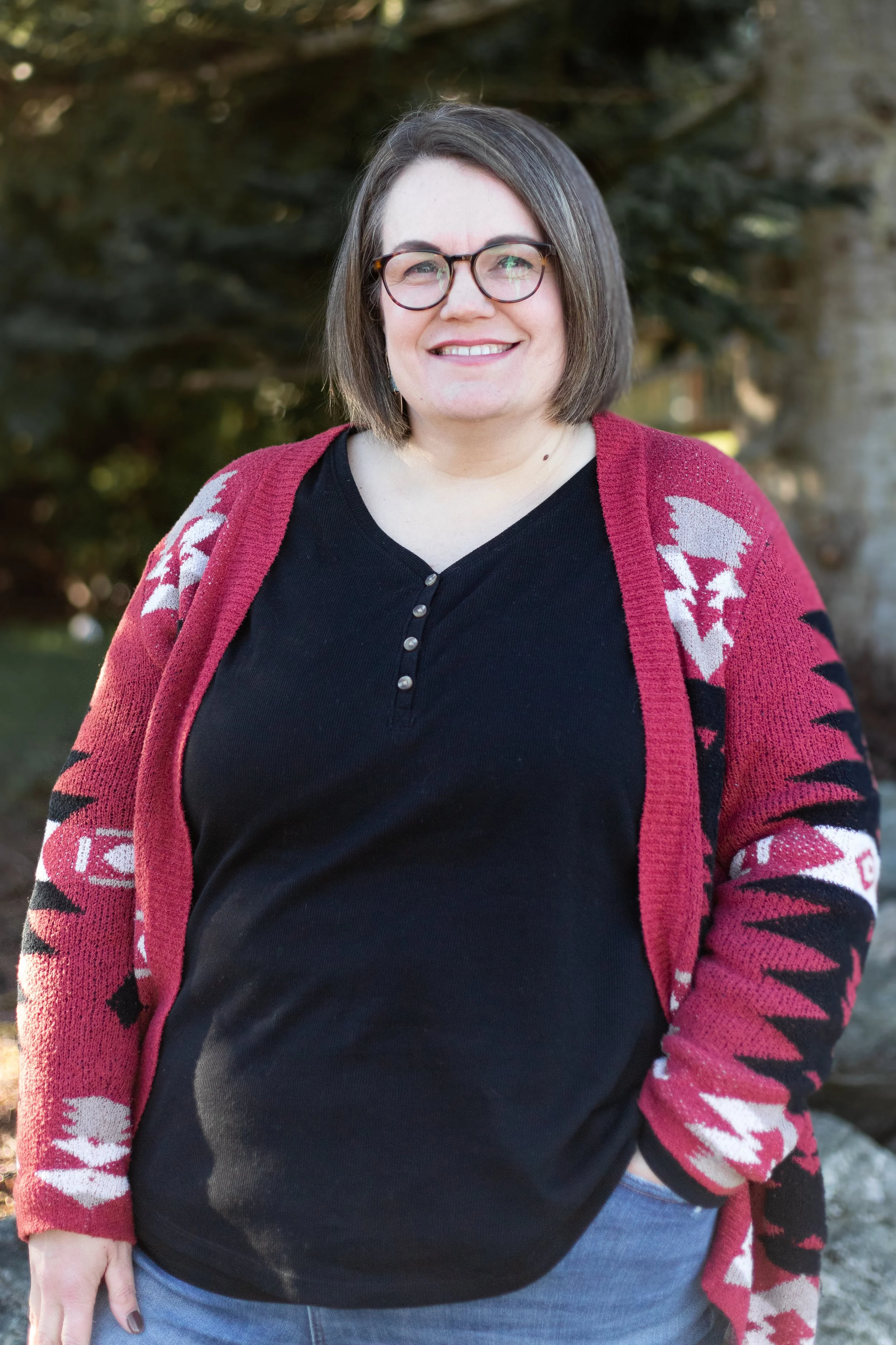 A woman with shoulder-length brown hair and glasses smiling outdoors, wearing a black shirt and a red patterned cardigan.