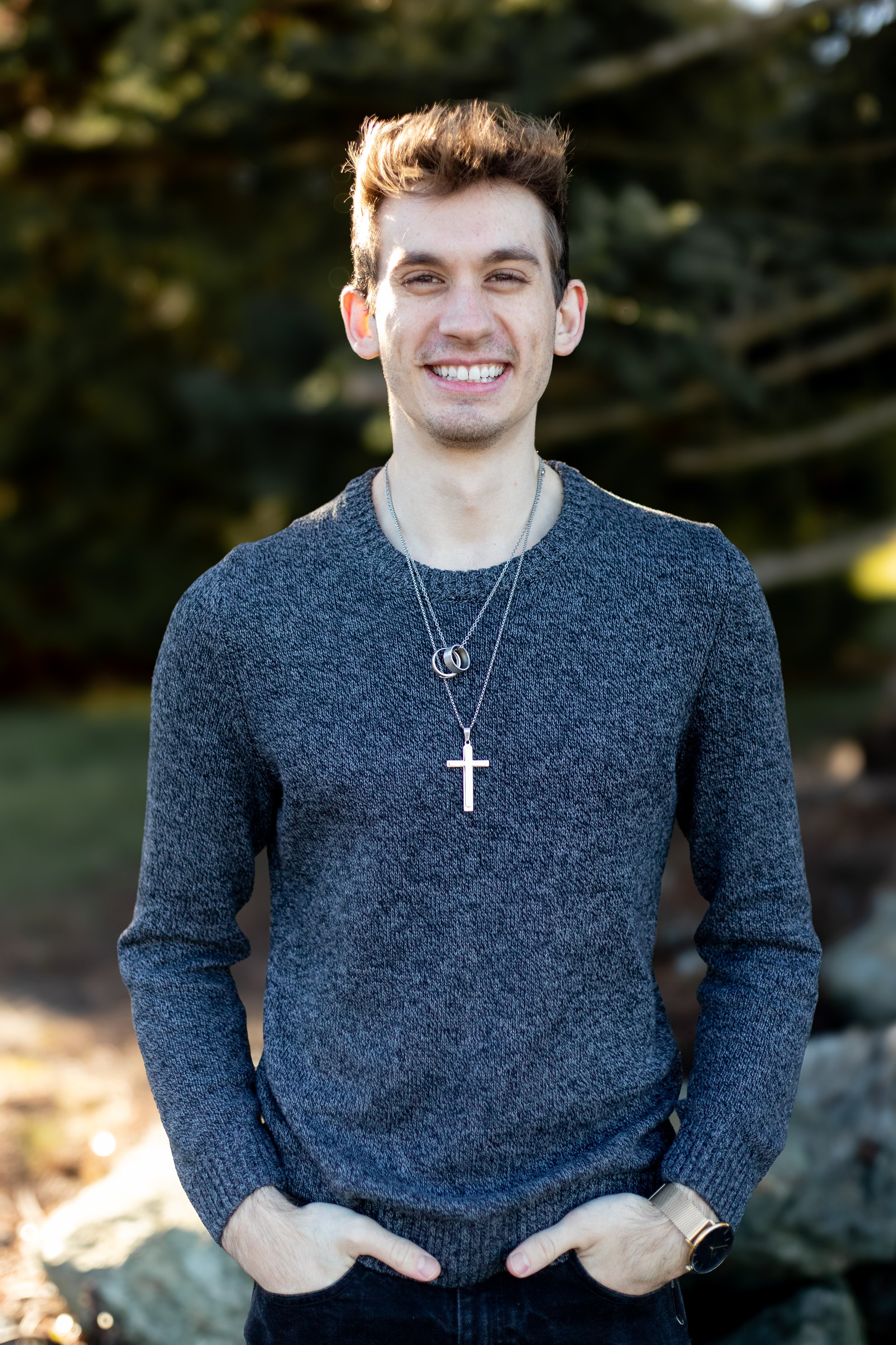 A smiling young man with light brown hair, wearing a gray sweater, layered necklaces with a cross and rings, standing outdoors in natural light.