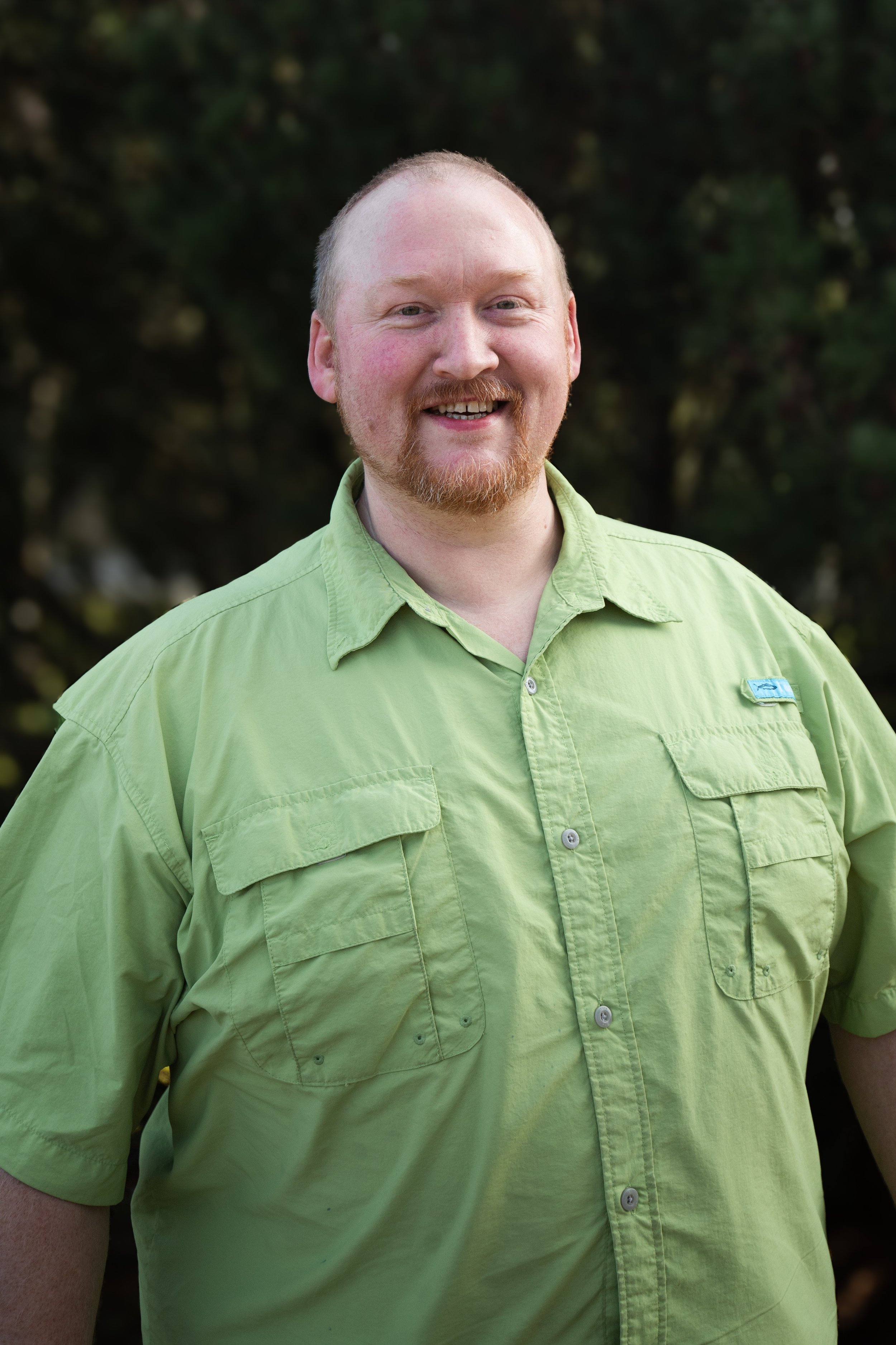 A smiling man with a beard in a light green button-up shirt standing outdoors with a blurry green background.