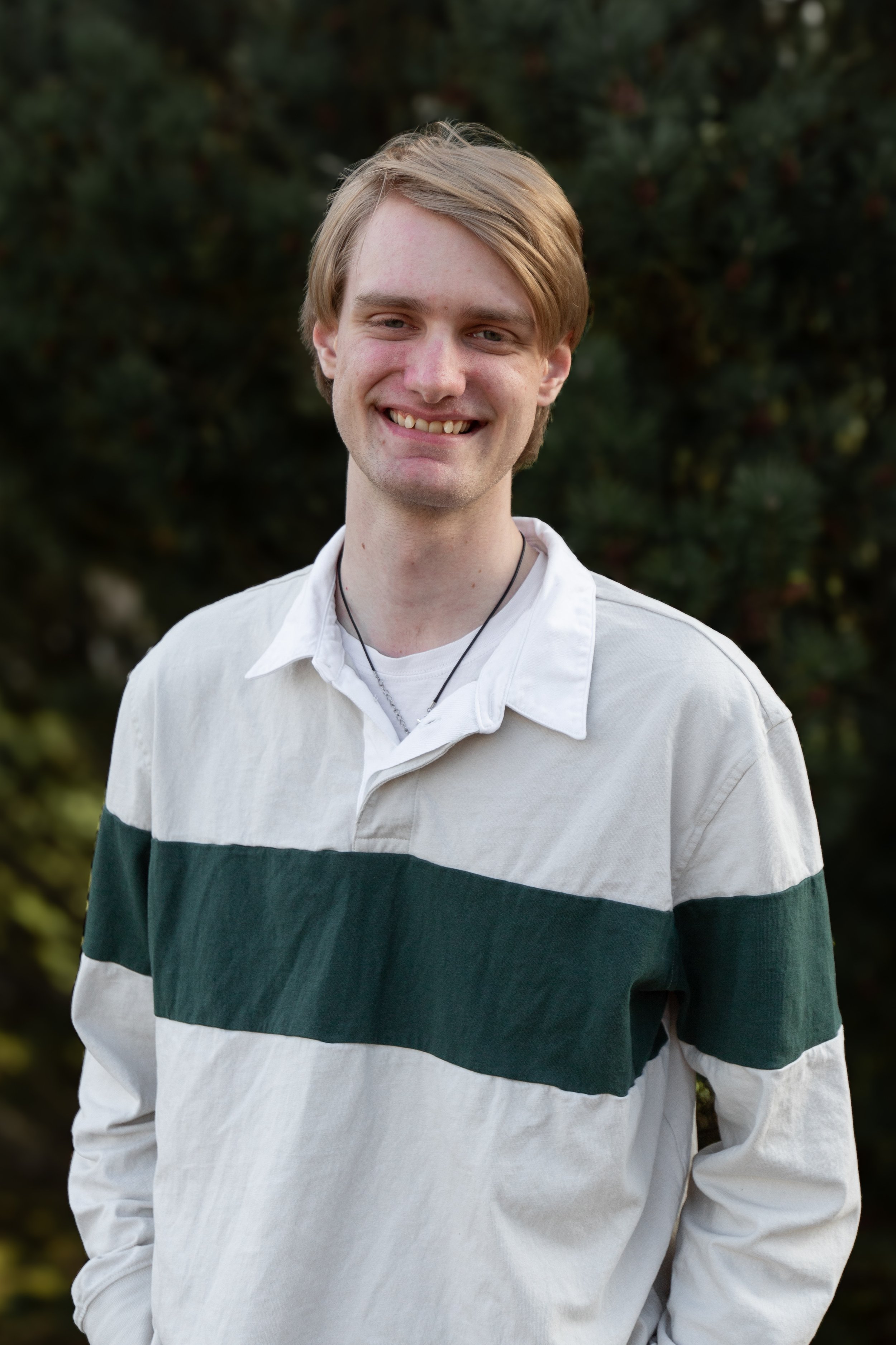 A young man with light skin and short, light brown hair, smiling outdoors. He is wearing a white and green striped long-sleeve shirt and a thin black necklace. The background is blurred greenery.