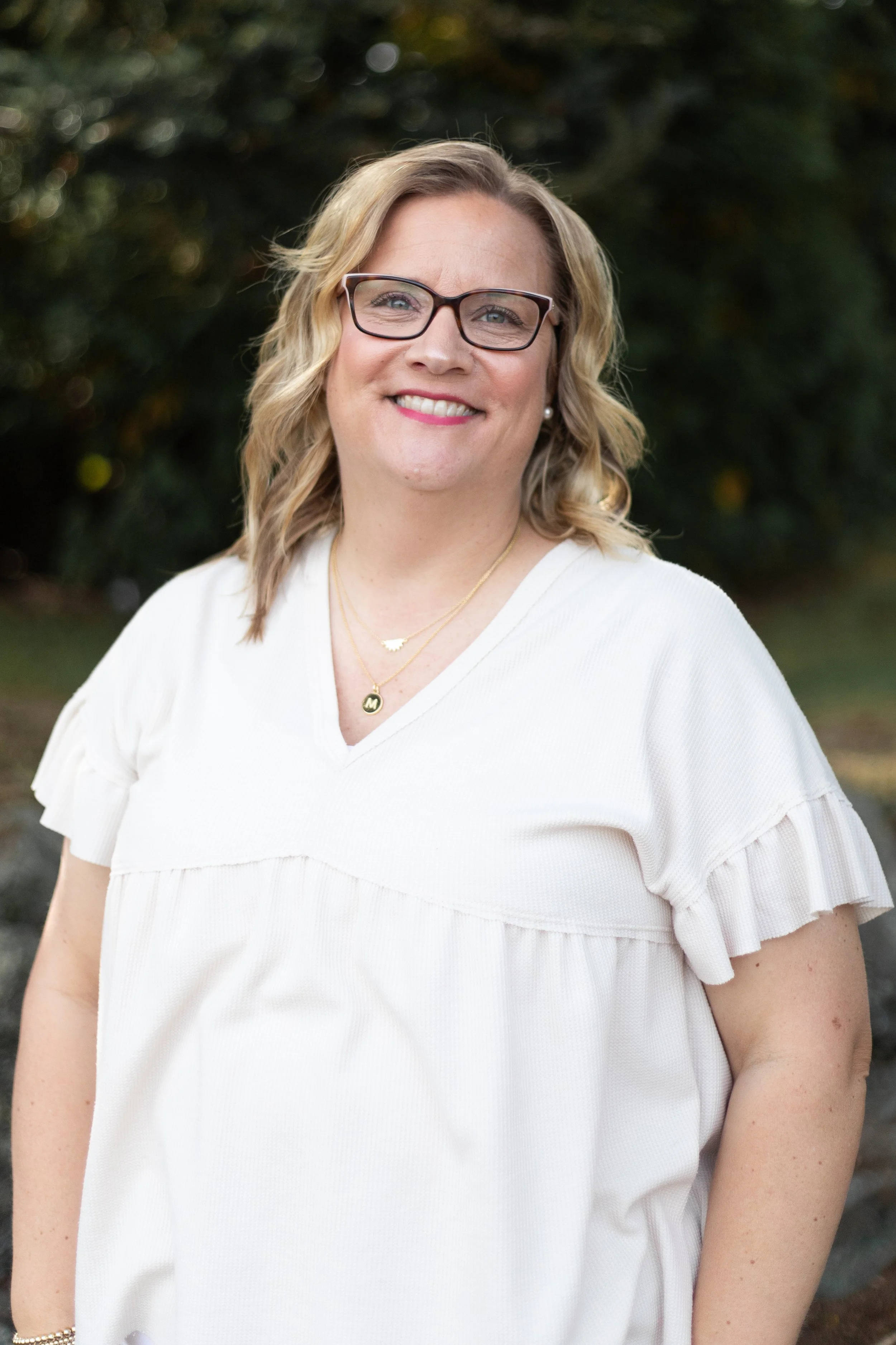 A woman with blonde hair, glasses, and a white top, standing outdoors in front of green foliage, smiling at the camera.