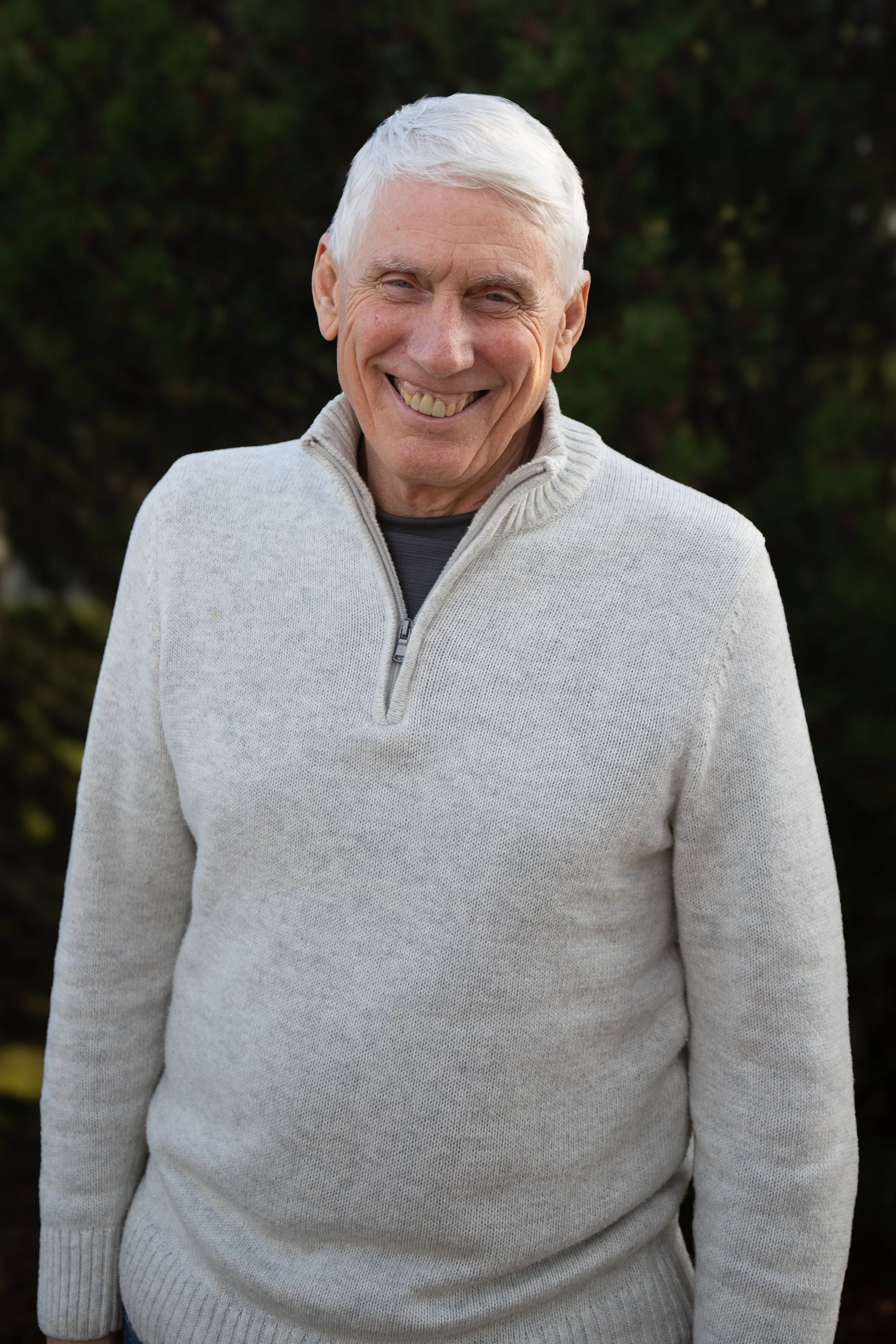 Smiling elderly man with white hair wearing a light gray zip-up sweater outdoors.