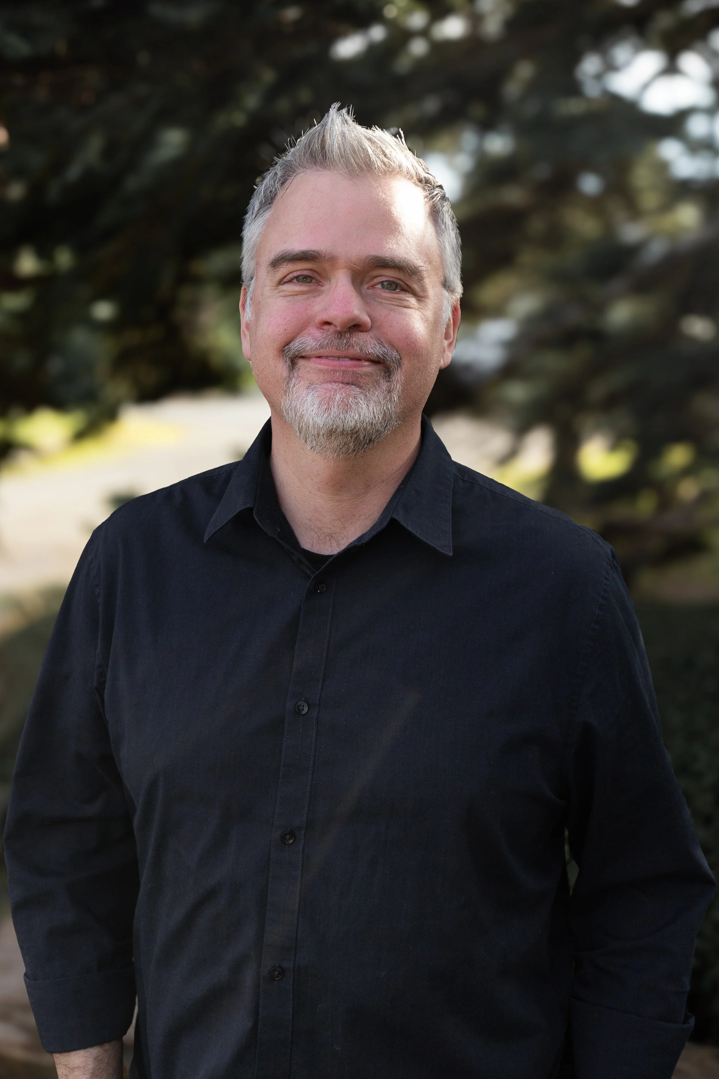 A middle-aged man with gray hair and a beard, wearing a black shirt, smiling outdoors with trees in the background.