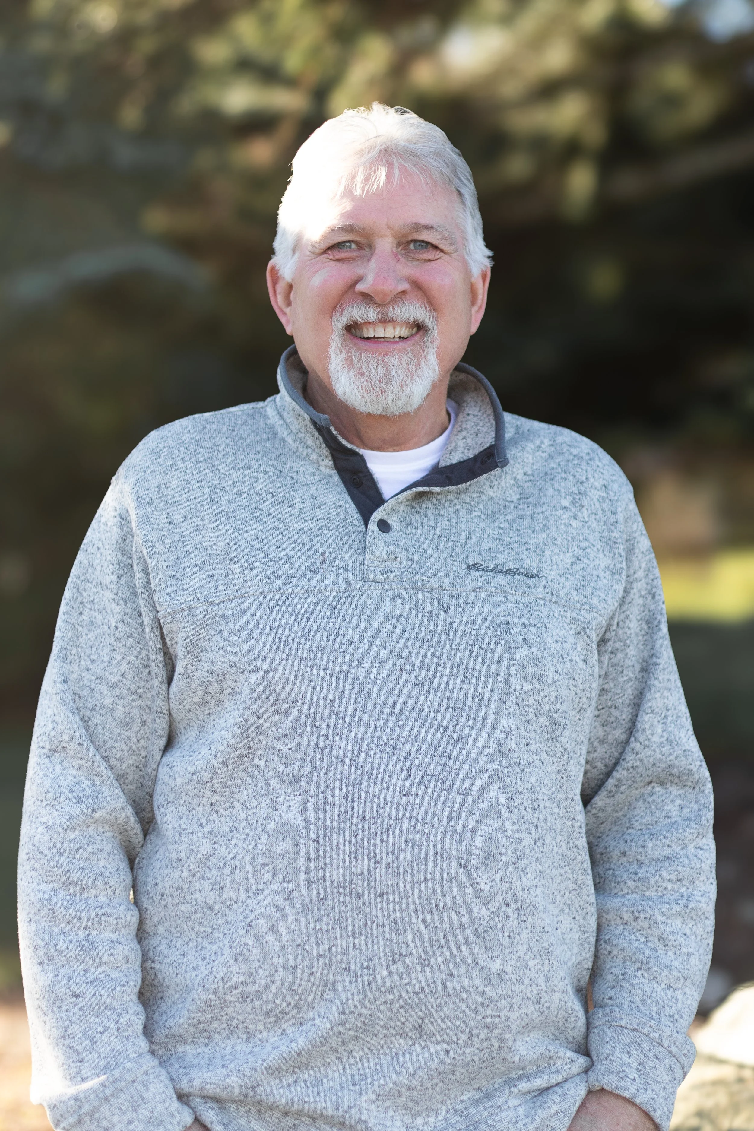 A smiling older man with white hair and a beard, wearing a light gray quarter-zip sweater with a black collar, standing outdoors with trees in the background.